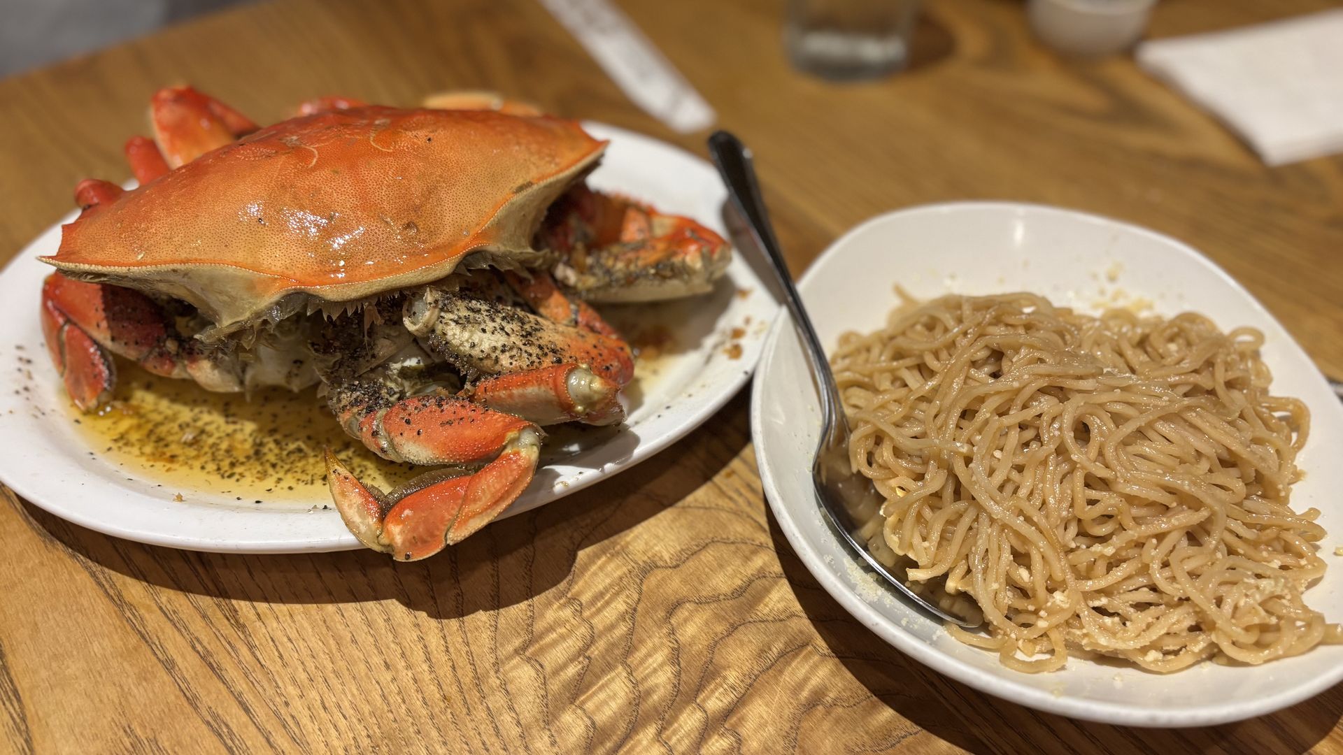 Cooked whole crab seasoned with black pepper and butter sauce on a white plate, served beside a white bowl of seasoned noodles with a spoon on a wooden table.