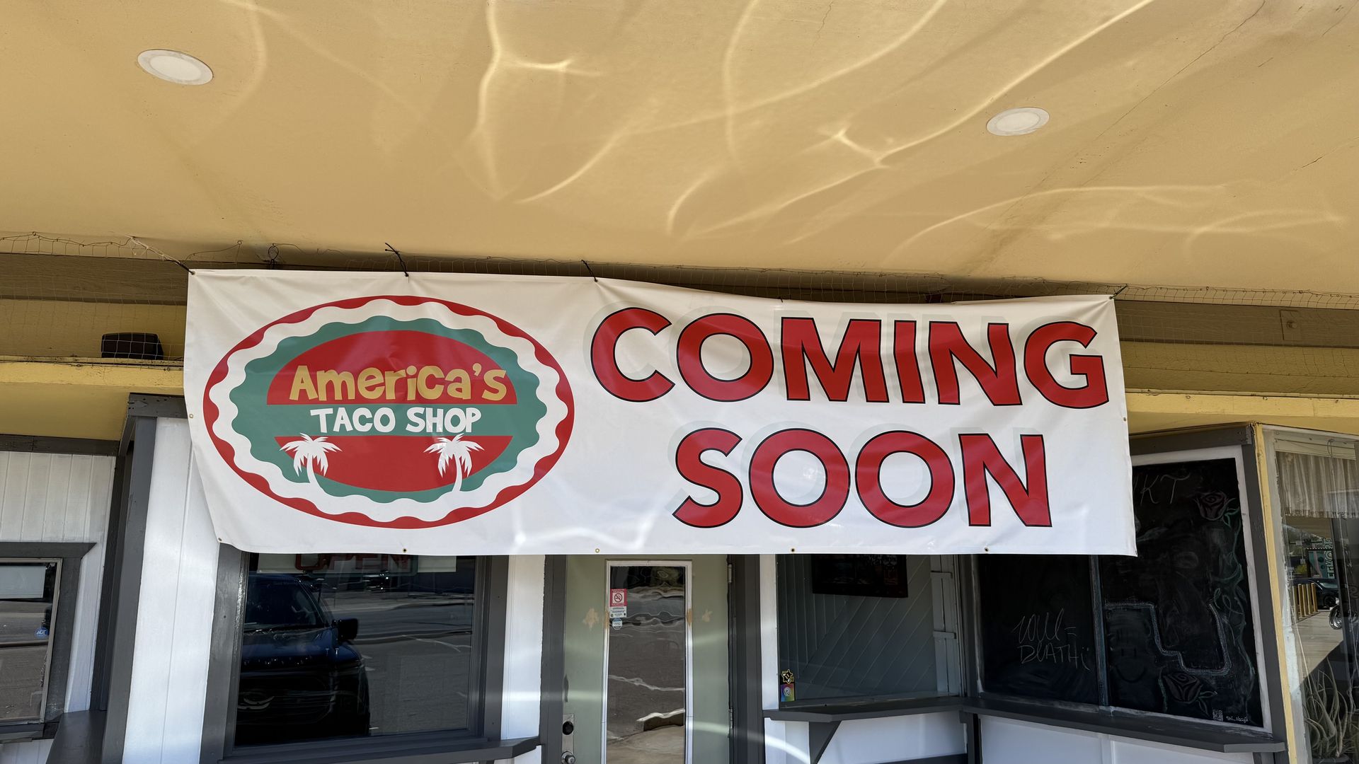Banner for "America's Taco Shop" shows a teal and red circular logo with palm trees, beside large red "COMING SOON" letters, hanging under a beige ceiling above storefront doors.