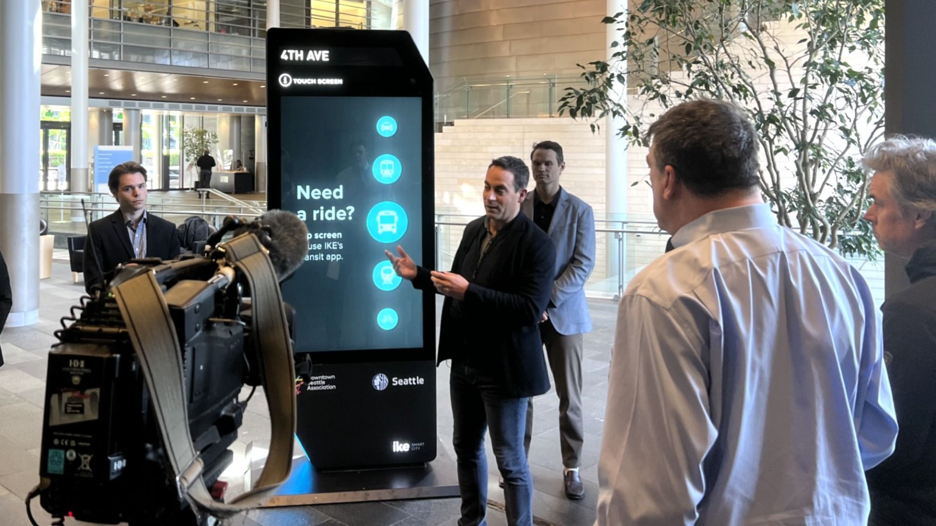 People gather around a digital interactive kiosk, which is several feet taller than the people around it, during a presentation at Seattle City Hall. A video camera is in the foreground.
