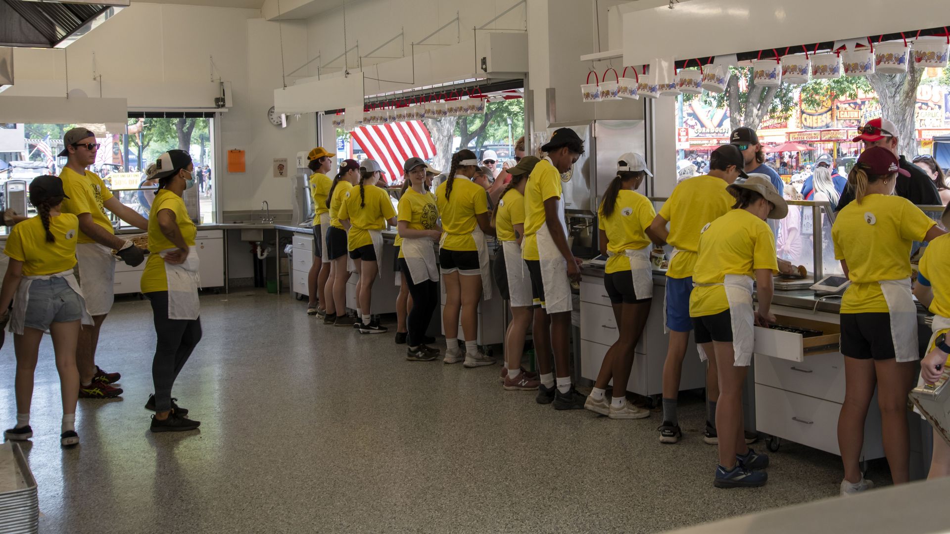 Bright festival concession stand with a long line of volunteers in yellow shirts and aprons at the counter. Kitchen area on the left; outdoor stalls and trees visible outside.