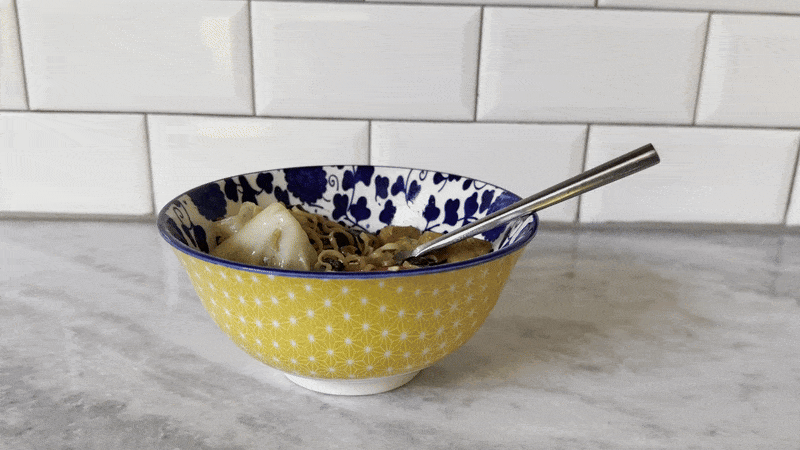 A blue and yellow bowl sits on a countertop while a hand reaches in and pulls a food laden with noodles and vegetables out of the bowl.