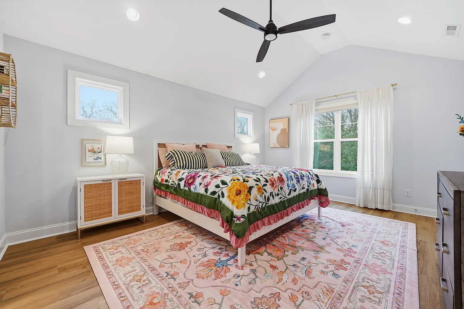 Bright bedroom with light gray walls, wooden floor, floral-patterned bedspread with yellow and pink flowers, pink area rug, black ceiling fan, white curtains, and natural light from three windows.
