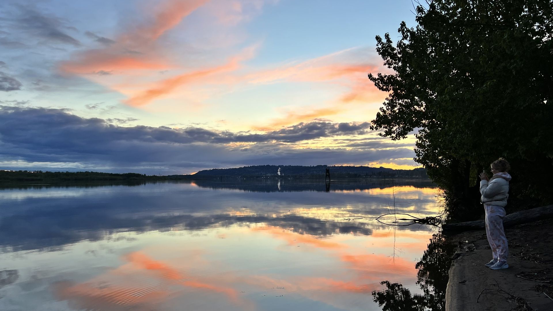 A person in a white jacket and patterned pants stands by a tree-lined riverbank during sunset, with a colorful sky of blue, orange, and pink clouds reflected on the calm water.