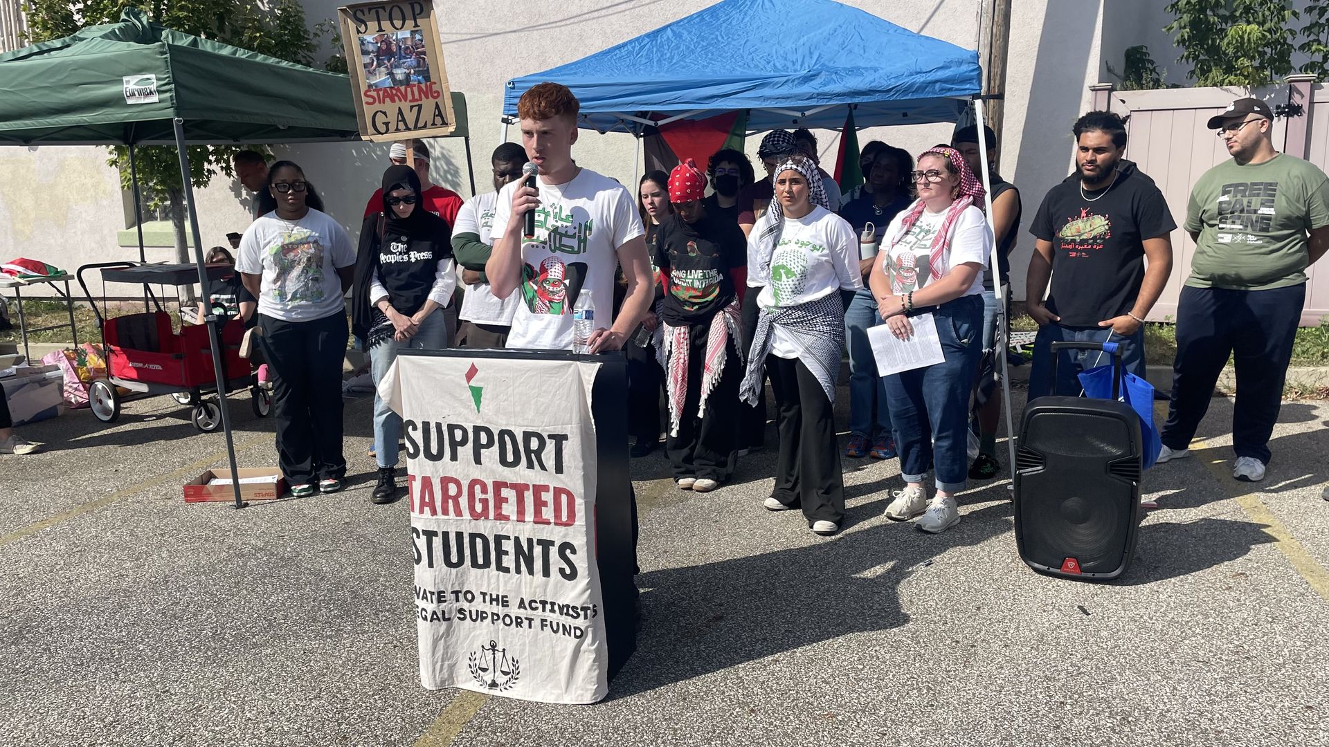 Group of diverse people standing outdoors under green and blue tents. One person speaks at a podium with a banner reading "Support Targeted Students." A sign says "Stop Starving Gaza."