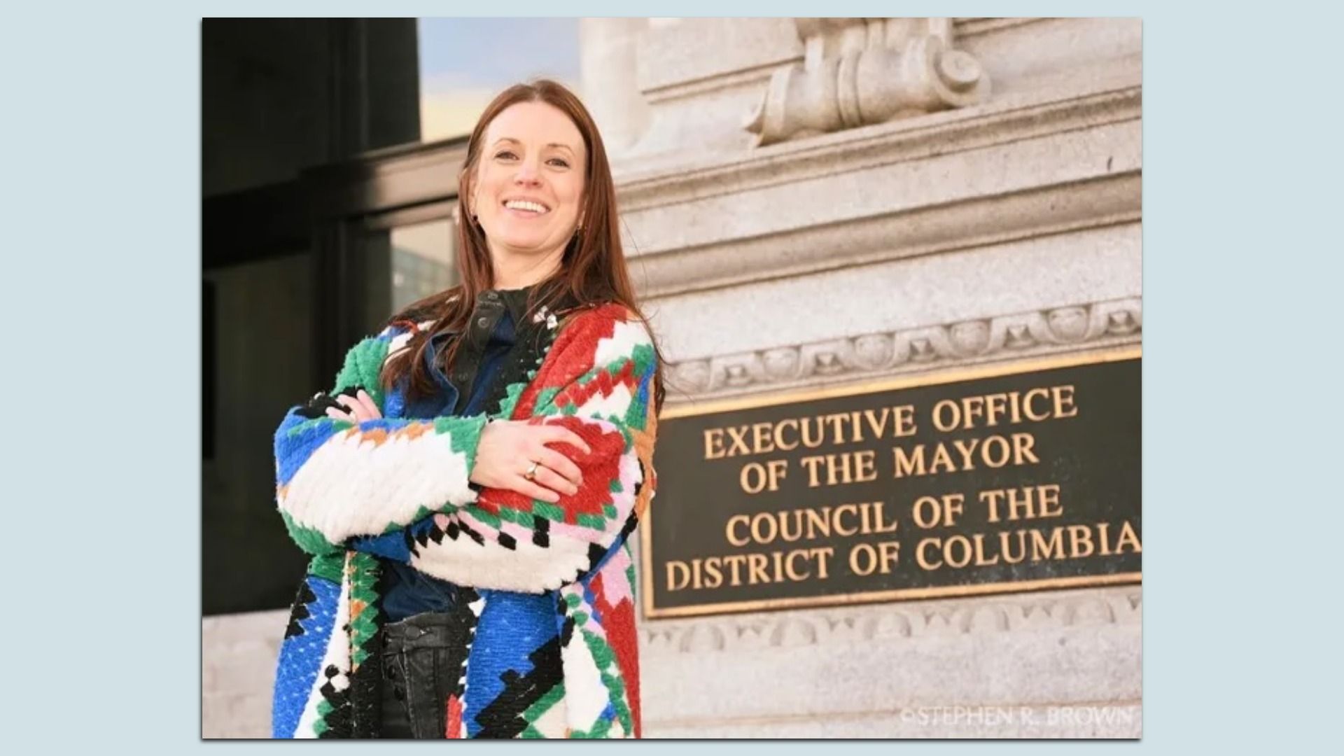 Hope Solomon photographed smiling with arms crossed wearing a colorful patterned coat standing in front of city hall with a sign reading "Executive Office of the Mayor Council of the District of Columbia".