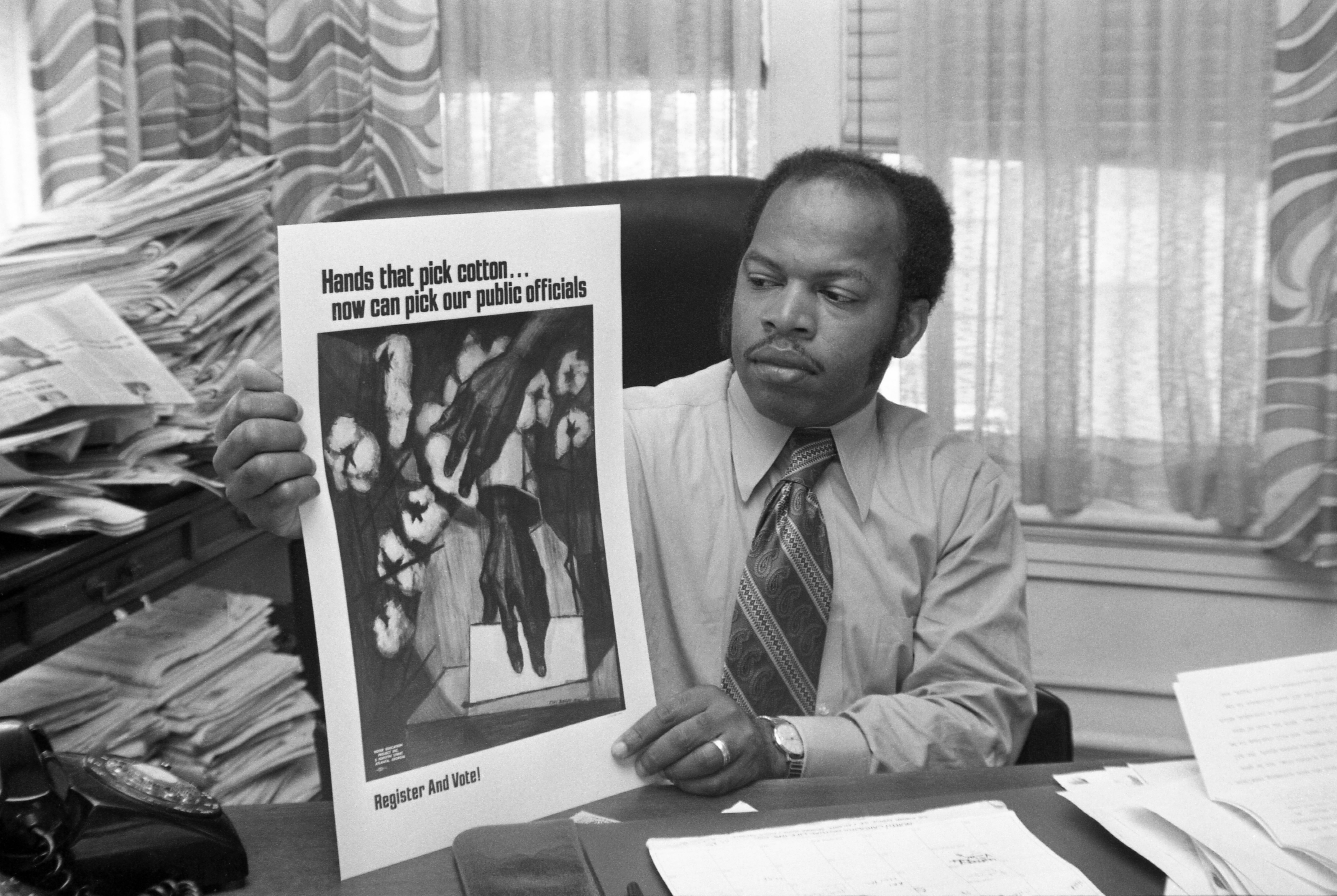 View of American Civil Right leader (and future US Representative) director of the Voter Education Project (VEP) John Lewis (1940 - 2020) as he sits at his desk and holds up a voter registration poster, Atlanta, Georgia, October 10, 1971. The poster reads 'Hands That Pick Cotton Can Now Pick Our Public Officials; Register and Vote.' (Photo by Bettmann Archive/Getty Images)