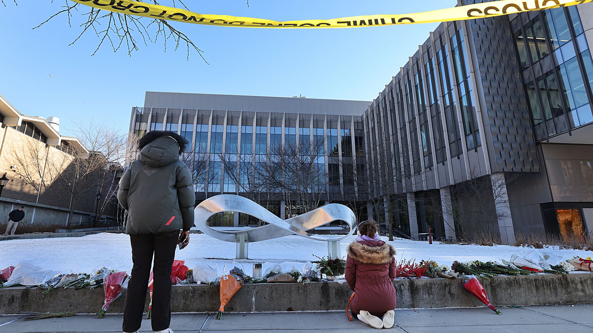 The back of a standing Brown University student wearing a large, gray winter coat that covers their hair and brown pants and another knealing in a maroon winter coat with faux fur trimming, exposing the white soles of her sneakers, look at a makeshift floral memorial to the school's shooting victims