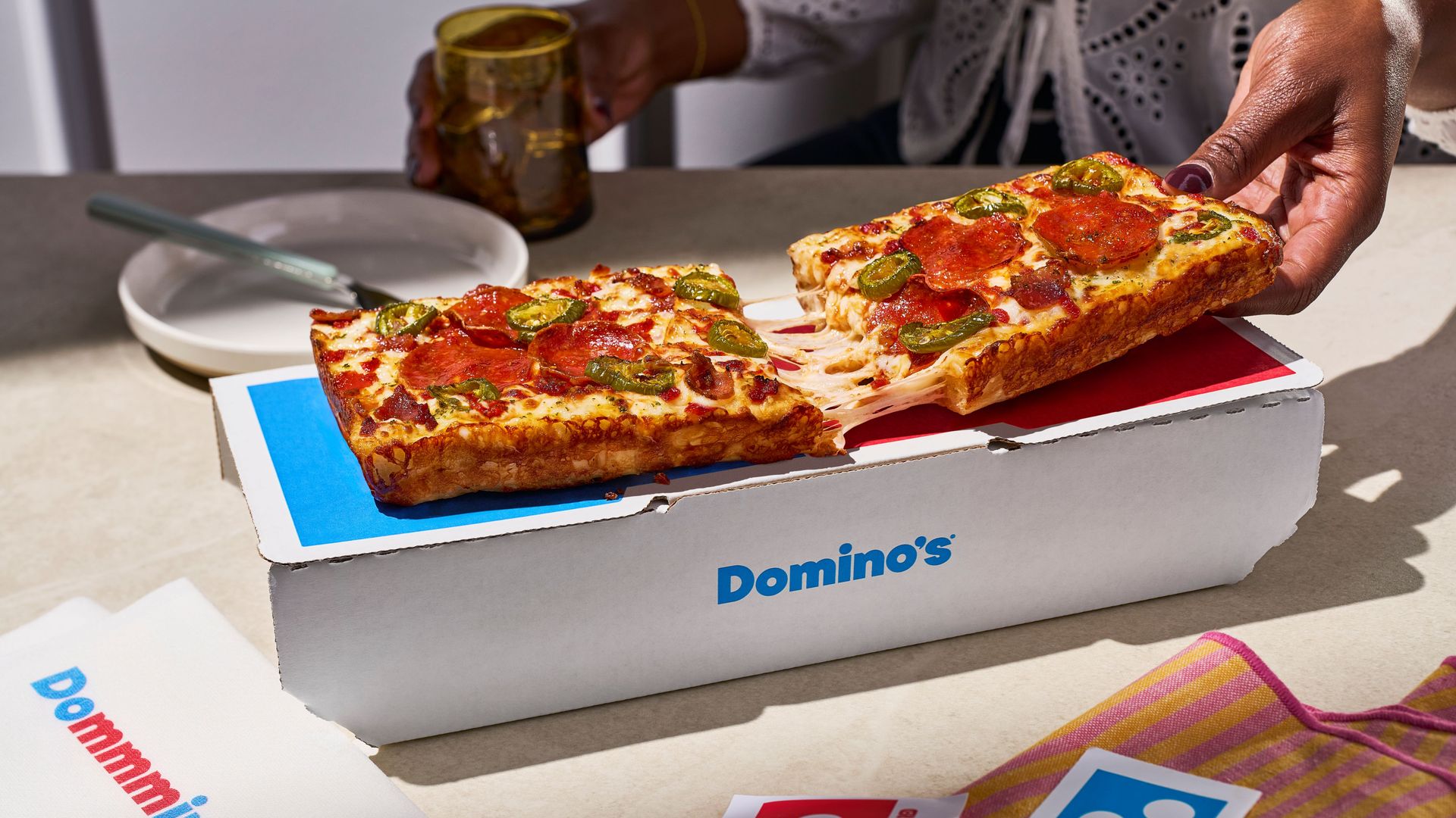 A Domino's pizza box sits open on a table outdoors as a hand pulls a slice of pepperoni and jalapeño cheese pizza; another slice remains. A bottle, plate, and a colorful napkin are nearby.