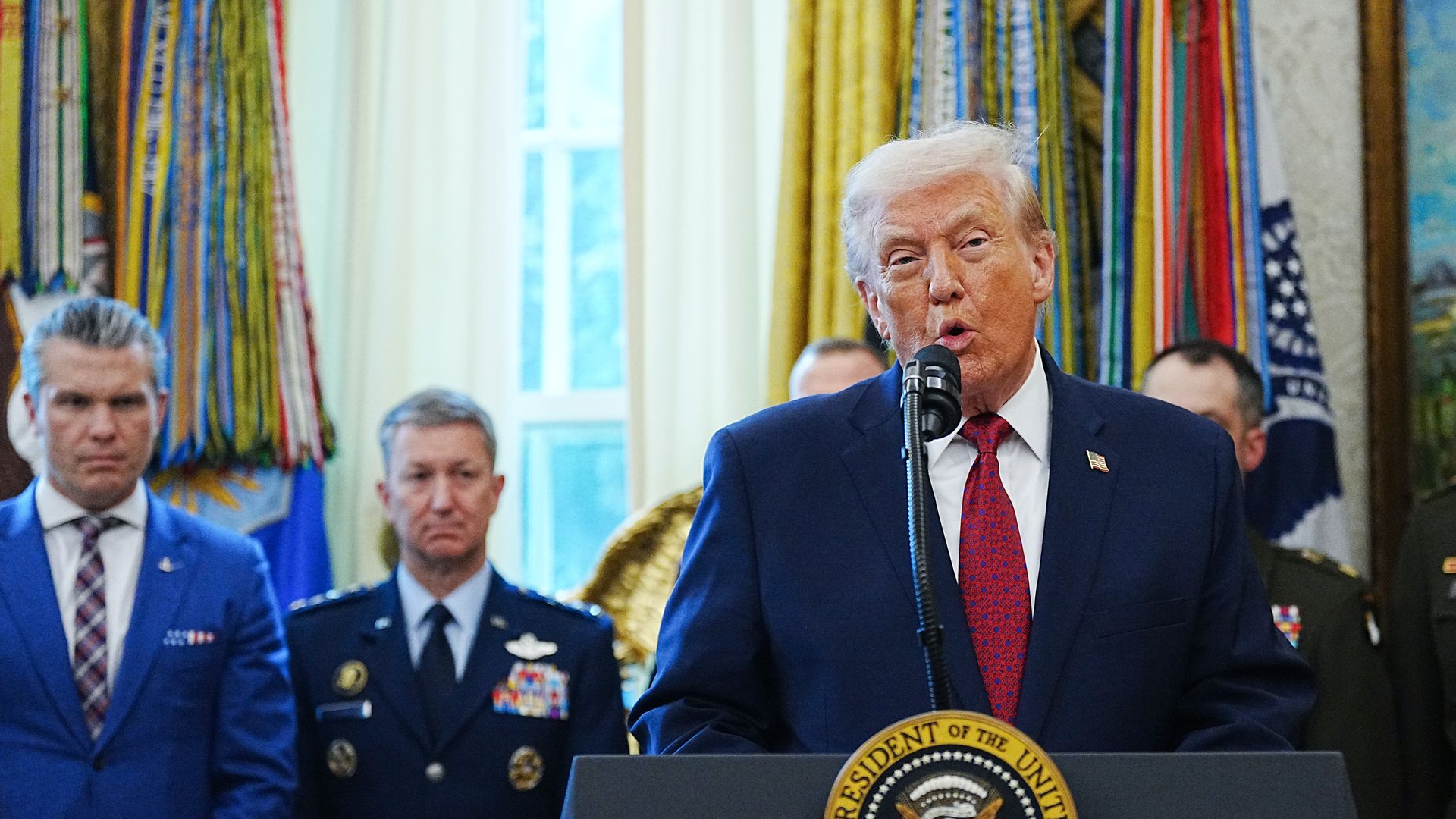 Trump wearing a blue suit jacket, white shirt and red and blue tie while speaking at a podium
