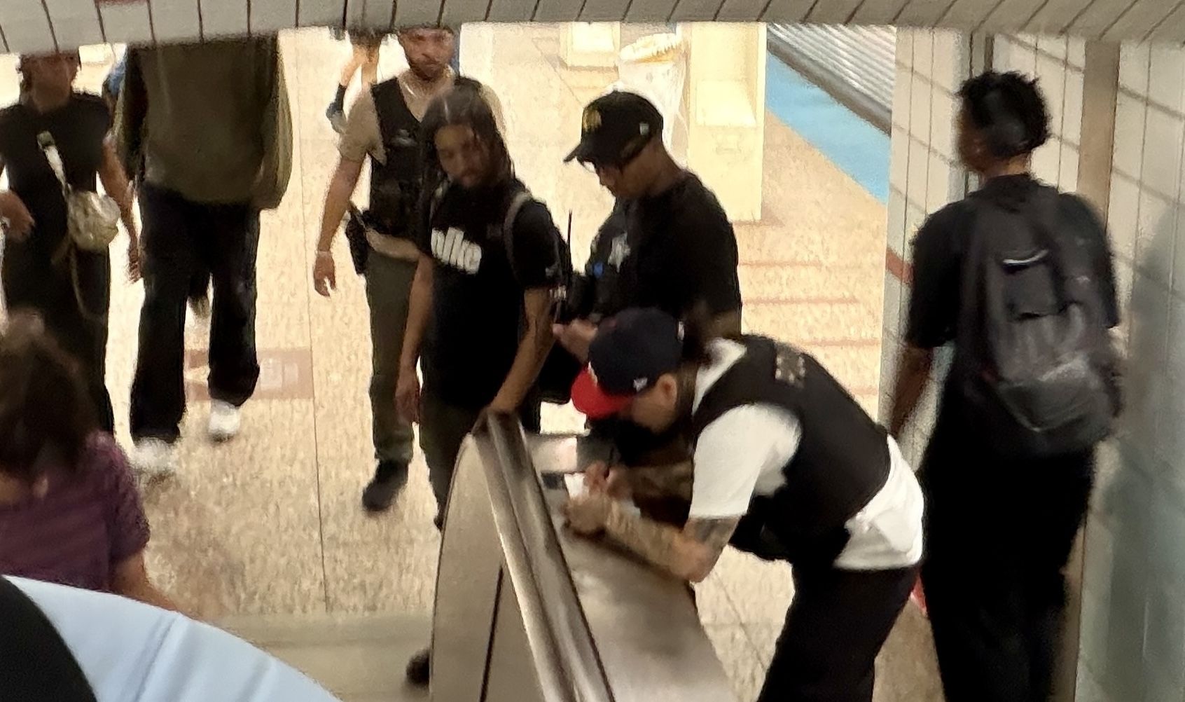 Group of people in a subway station around an escalator. A man wearing a black vest and red cap with a mask leans on the railing while others with backpacks stand nearby.
