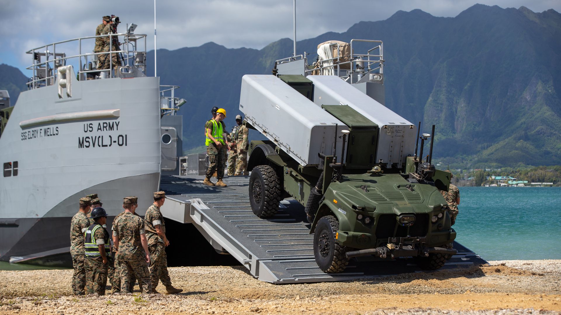 U.S. military vehicle unloading from gray ship near rocky shore with Marines in camouflage and helmets. Green mountains and blue water are in the background.