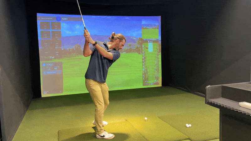 Gif shows a loop of Axios reporter Travis swinging a golf club and hitting a ball off of a tee into a screen as part of a golf simulator.