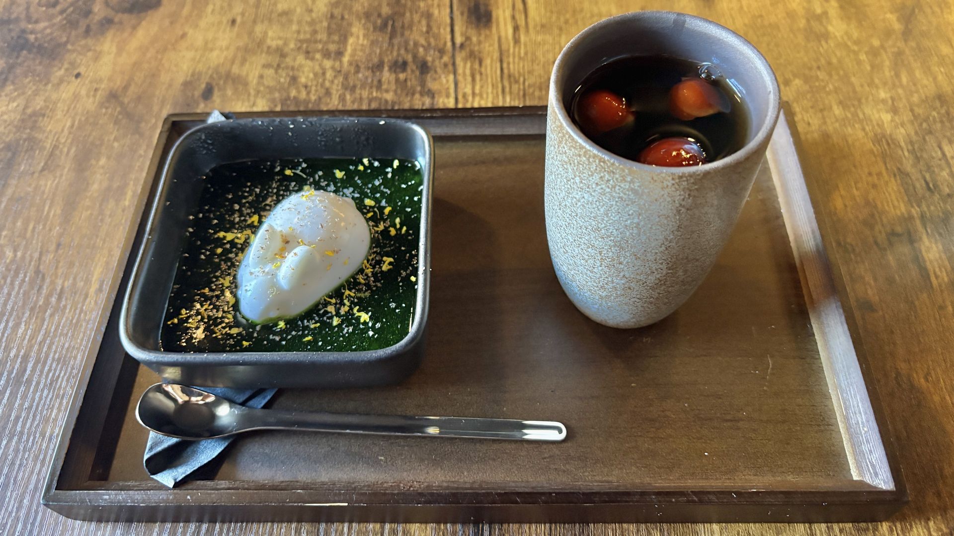 A wooden table with a tray: left, a square black dish of green jelly topped with a white scoop and orange zest; right, a tall, speckled ceramic cup with a dark drink and floating red berries.