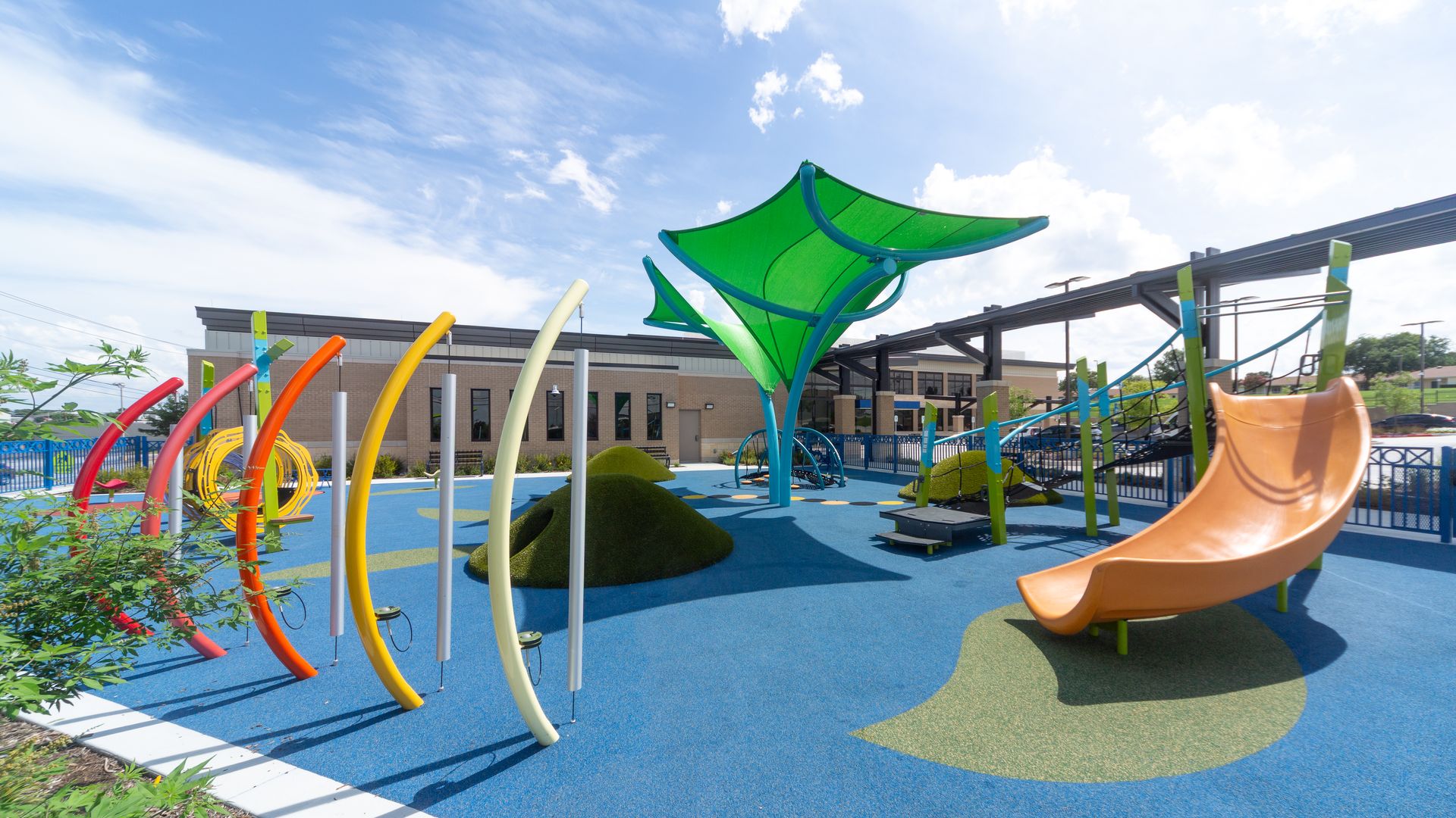 Bright, modern playground with blue and green flooring, colorful curved structures and chimes, a large green canopy, orange slide, and climbing features, under a partly cloudy sky.