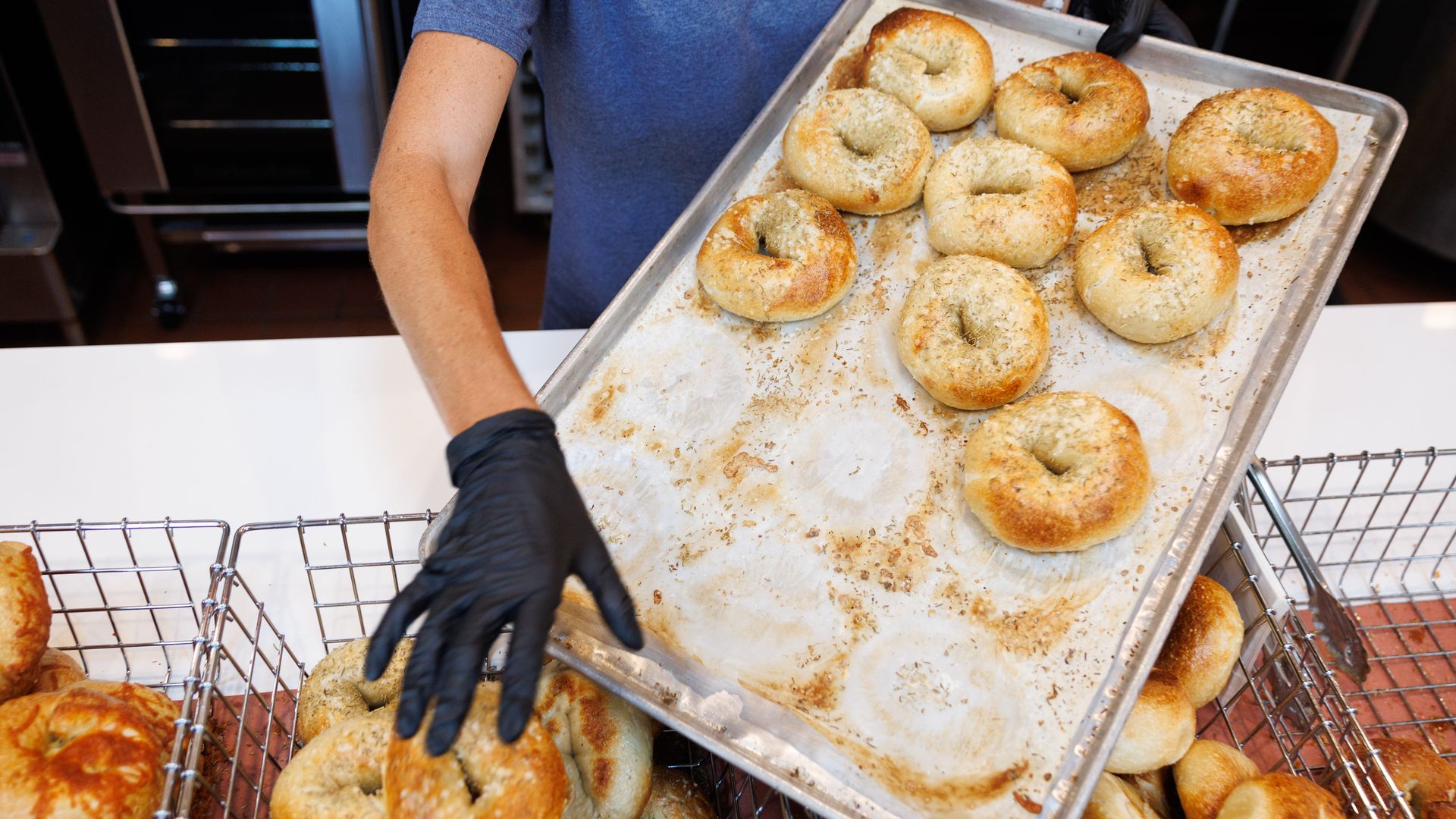 Person in a blue shirt and black gloves holds a baking tray of freshly baked bagels, sprinkled with sesame and salt, as racks and an oven appear in the background.