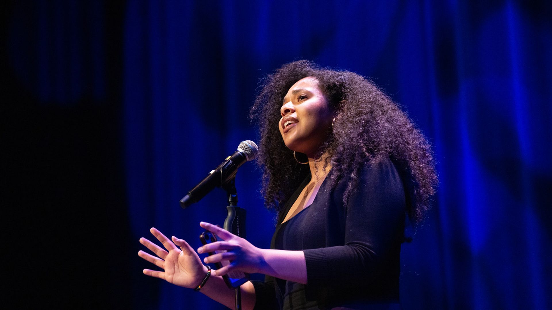 A young light-skinned black woman speaking at a mic in front of a blue velvet curtain