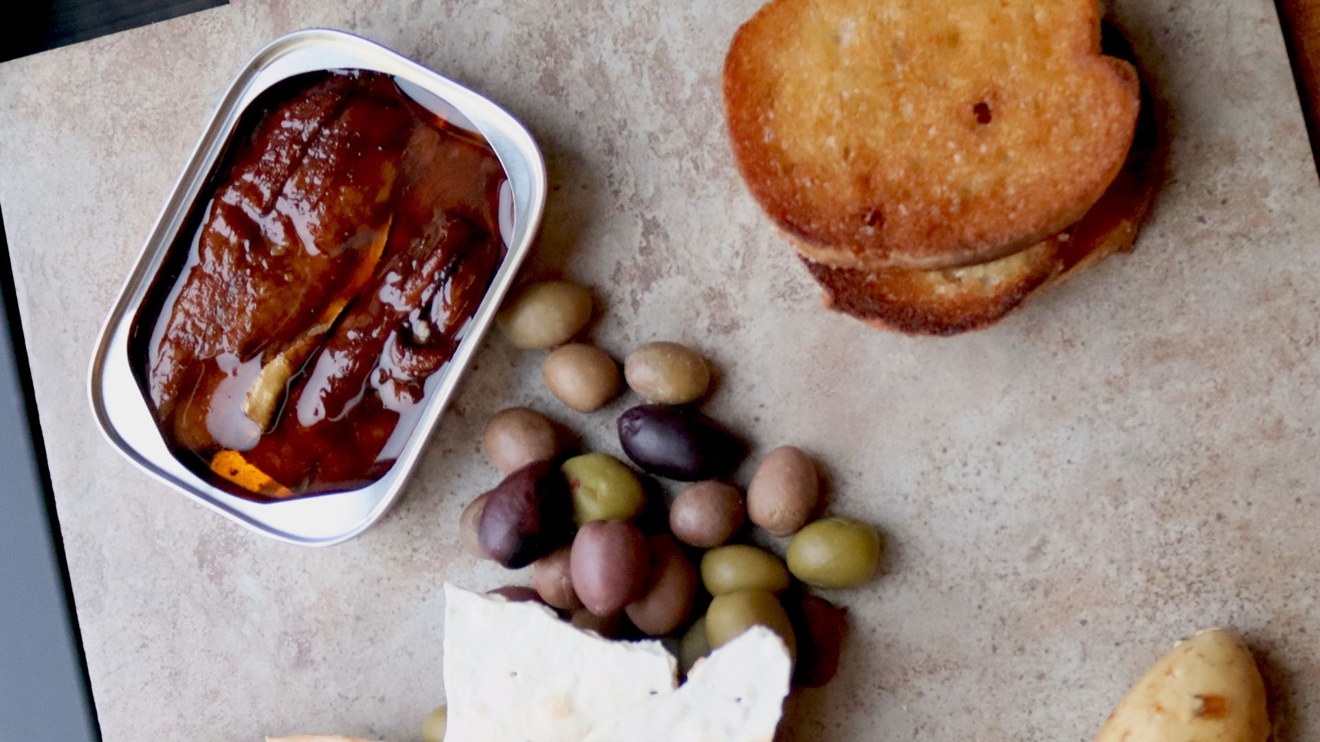 Open can of sardines in oil, assorted green and black olives, toasted bread slices, flatbread, and a small potato on a beige surface.