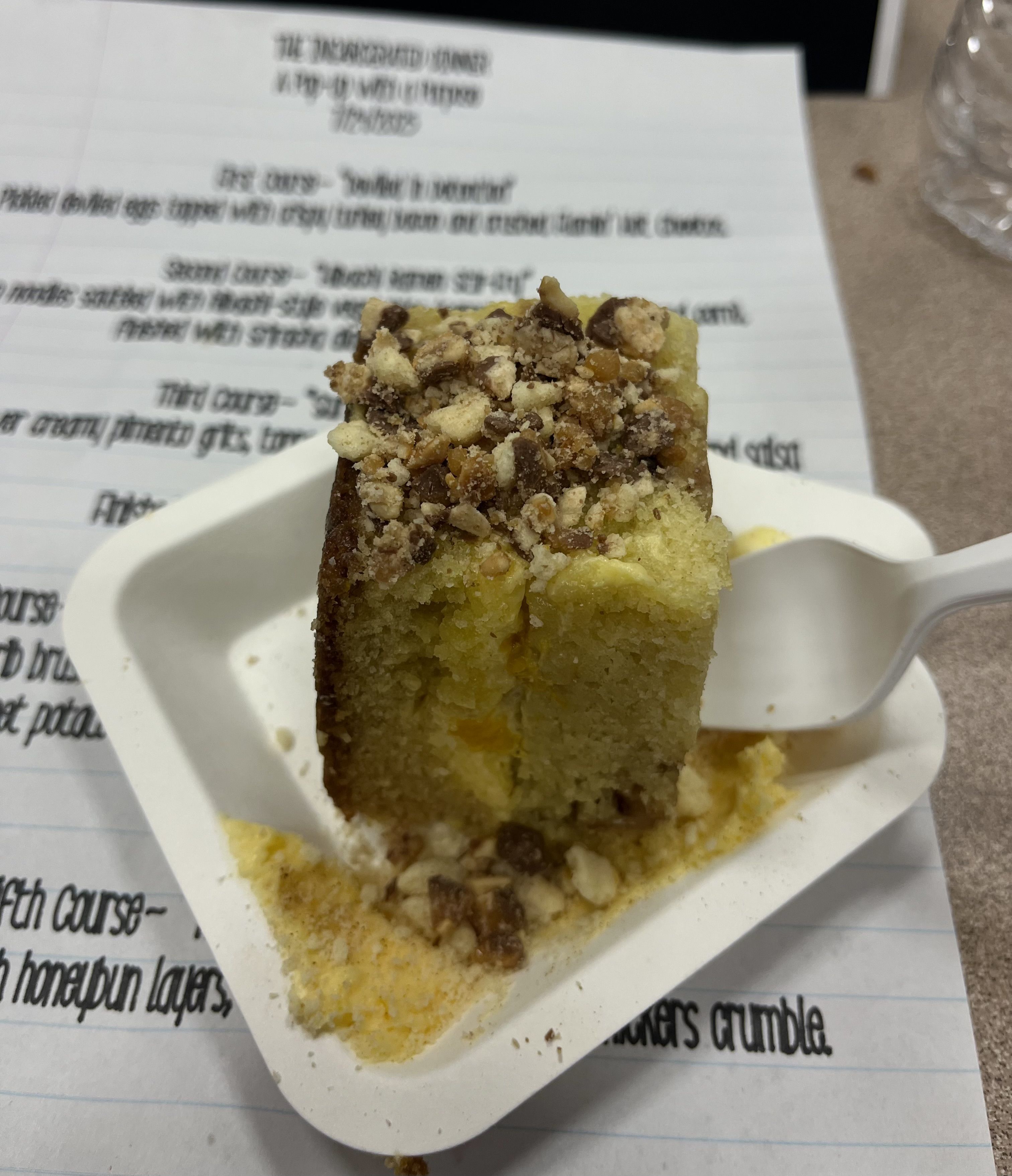 Close-up of a square slice of yellow cake with crumbly nut topping on a white plate with a white plastic spork, placed on a table with a typed menu sheet underneath.