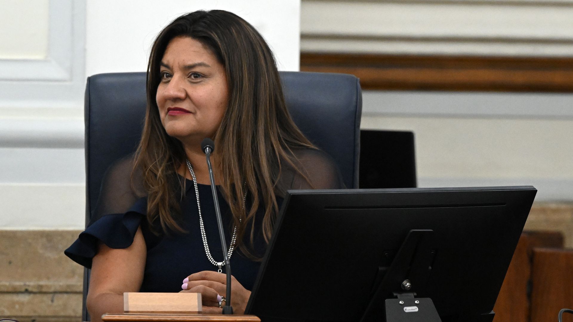 A woman with long dark hair and a pearl necklace sits at a dark wooden dais in a formal chamber, speaking into a microphone. A black nameplate reads ROMERO CAMPBELL in front, with wood paneling behind.