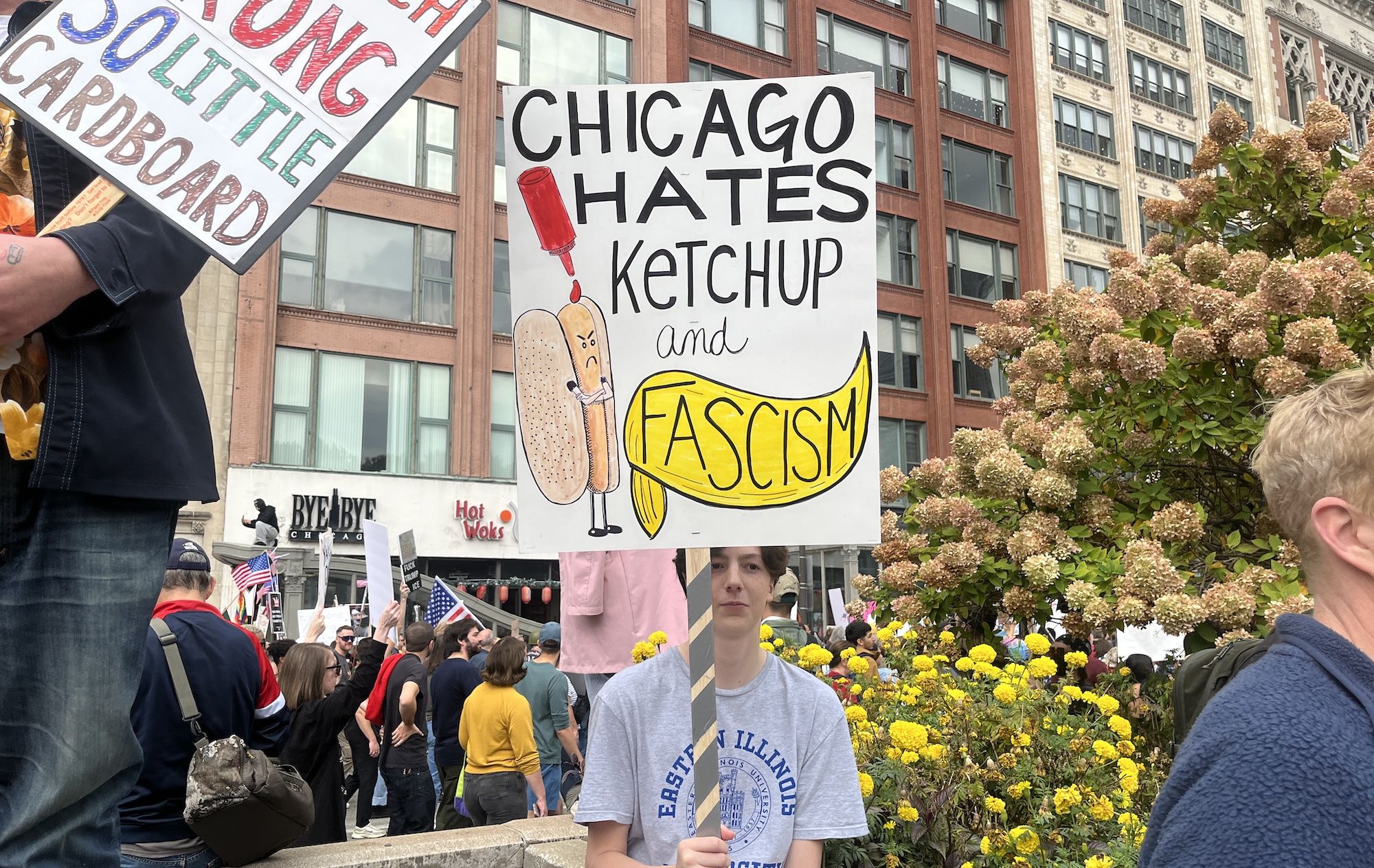 Jamie Nicholson-Creswell holding a protest sign reading "Chicago hates ketchup and fascism" with a drawing of an angry hotdog and a yellow banner. Crowd and buildings in background.