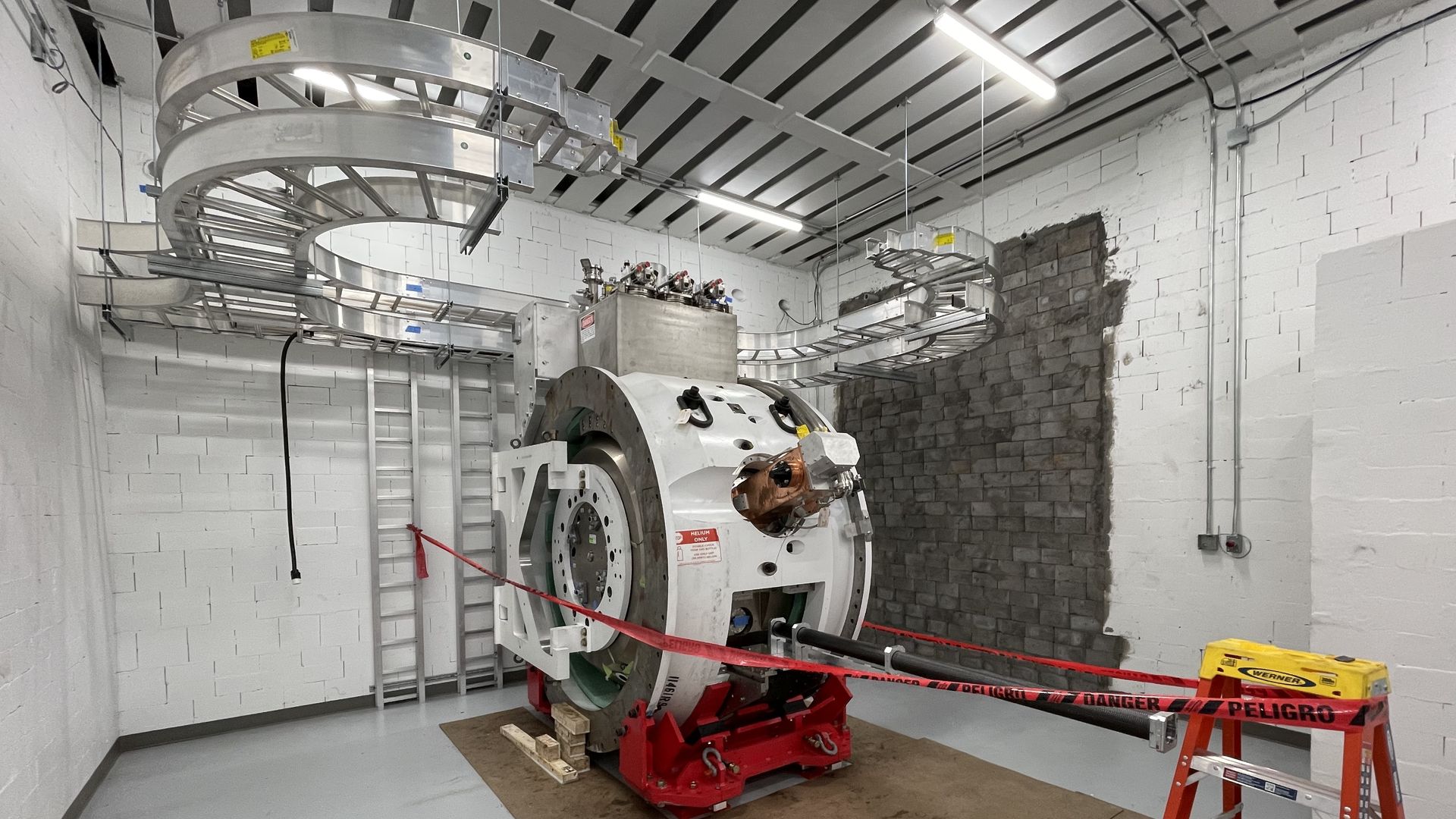 Industrial lab with a large circular metal frame/rotor, suspended rails overhead, a heavy machine, white brick walls, red caution tape, ladder, and exposed wiring under bright lights.