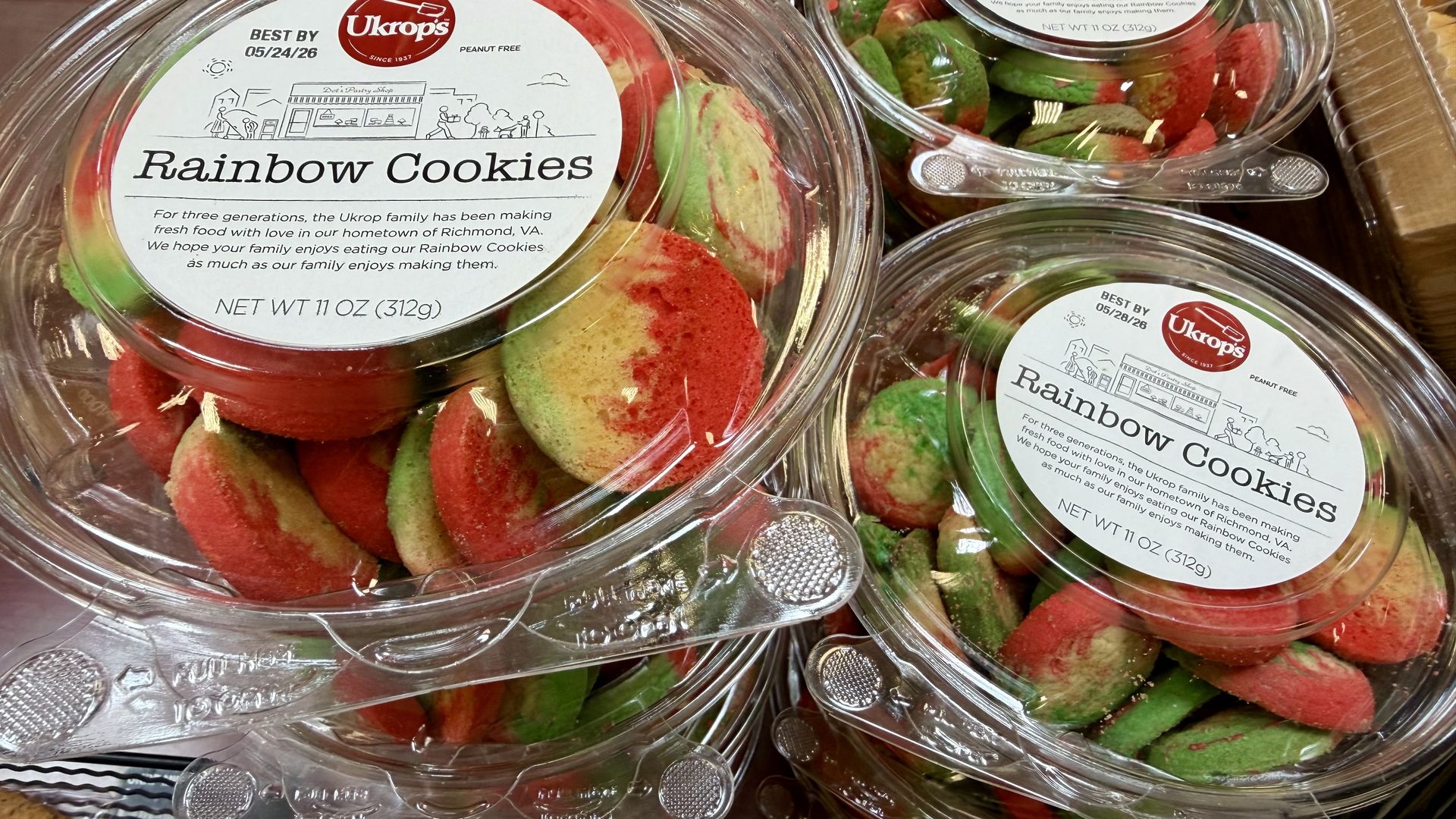 Close-up of clear plastic containers piled with red and green rainbow cookies. Labels read "Rainbow Cookies" from Ukrop's, showing a Best By date and net weight 11 oz (312 g).