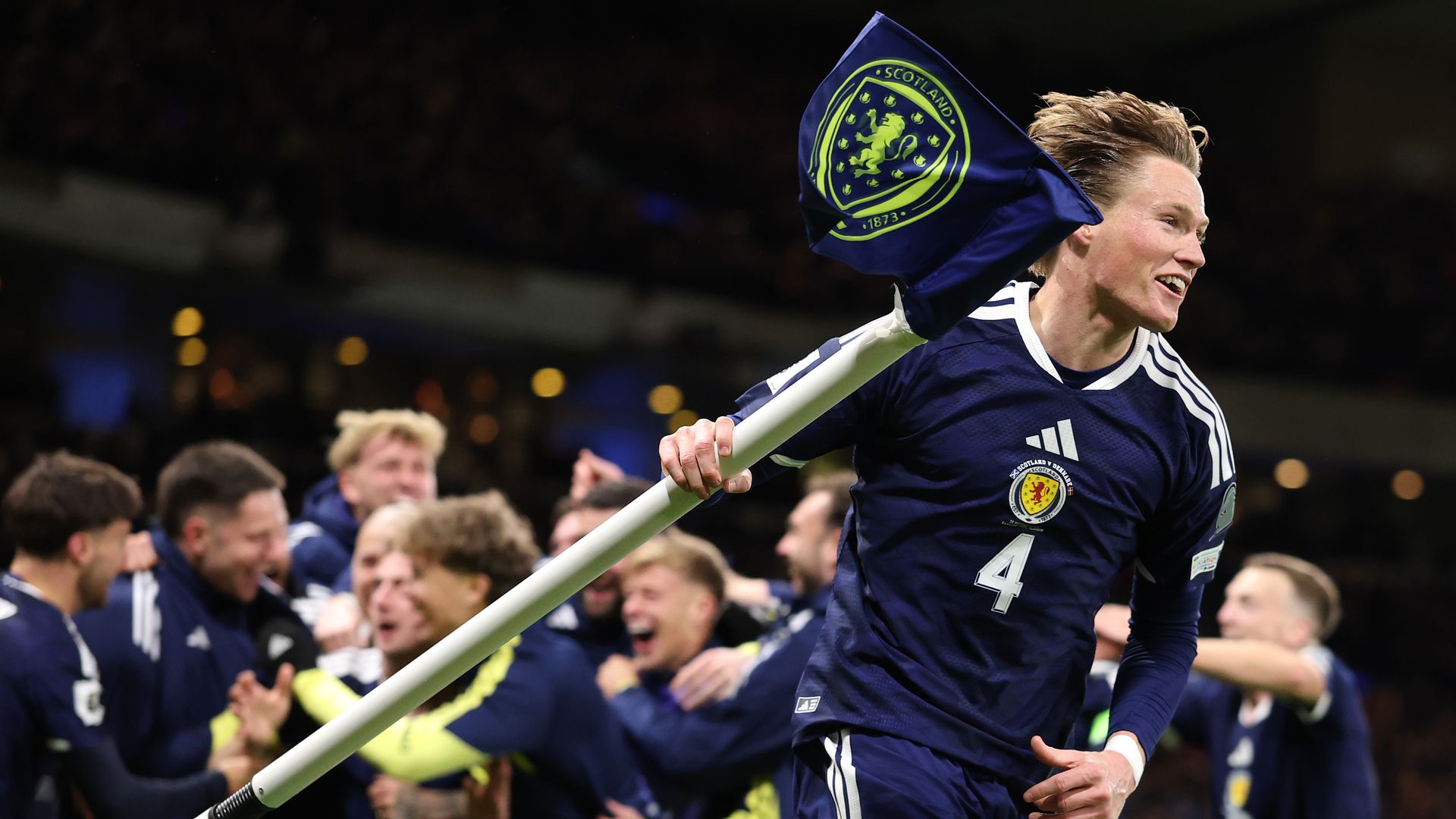 Scott McTominay of Scotland wearing dark blue jersey with number 4, celebrating with teammates in background, holding a corner flag with Scotland crest on stadium field at night.