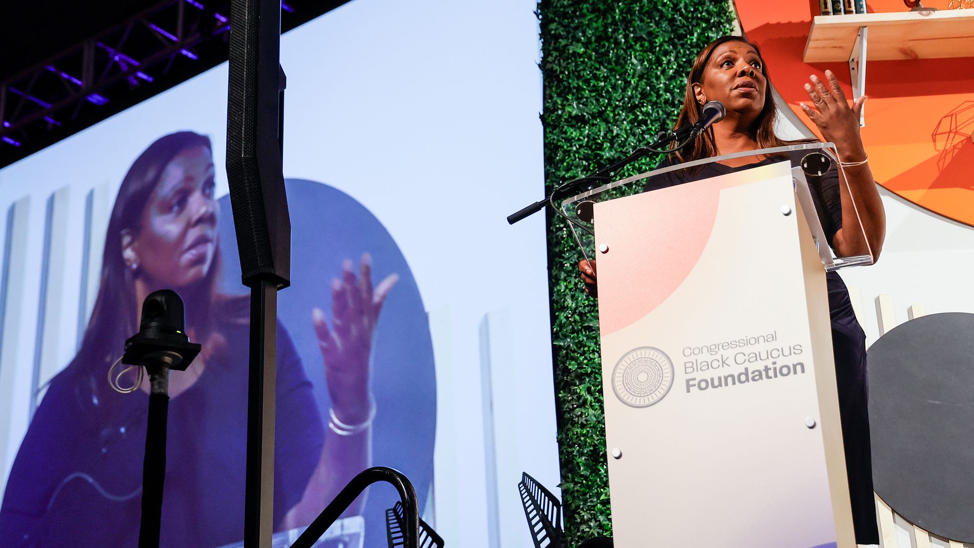 A woman speaks at a conference while an image of her speaking is projected behind her much larger. 