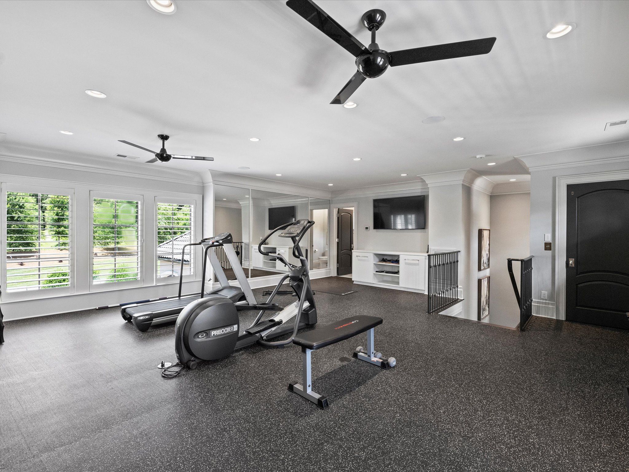 Bright home gym with black treadmill, elliptical, and bench on speckled black floor, large windows, mirrors, ceiling fans, and a wall-mounted TV in a white room with black doors.