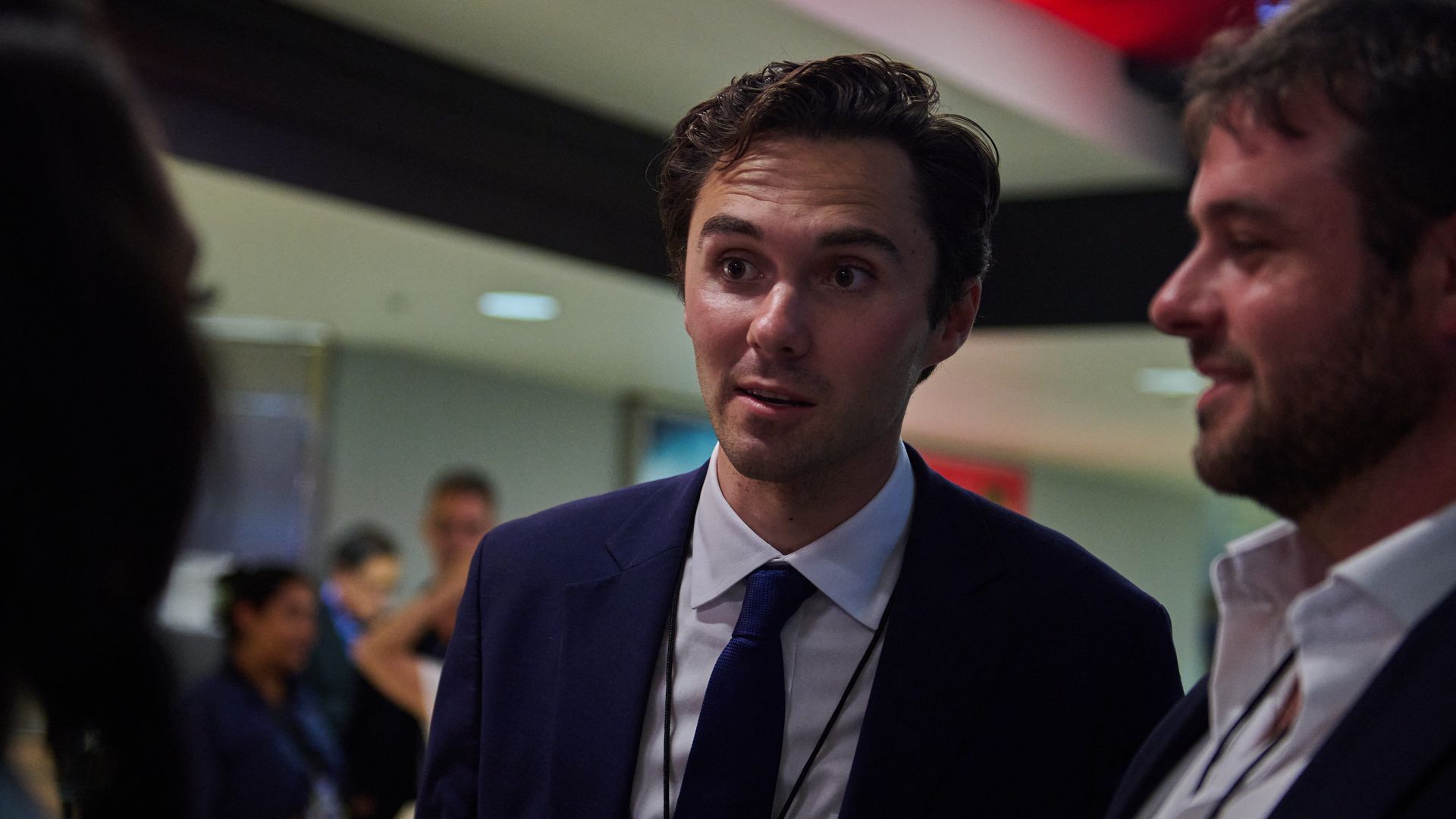 David Hogg, wearing a blue suit and tie and standing with others in a dimly lit hallway.