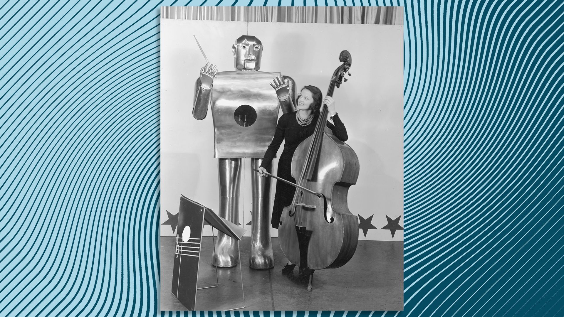 A midcentury photograph of a large robot standing next to a woman playing the cello. 