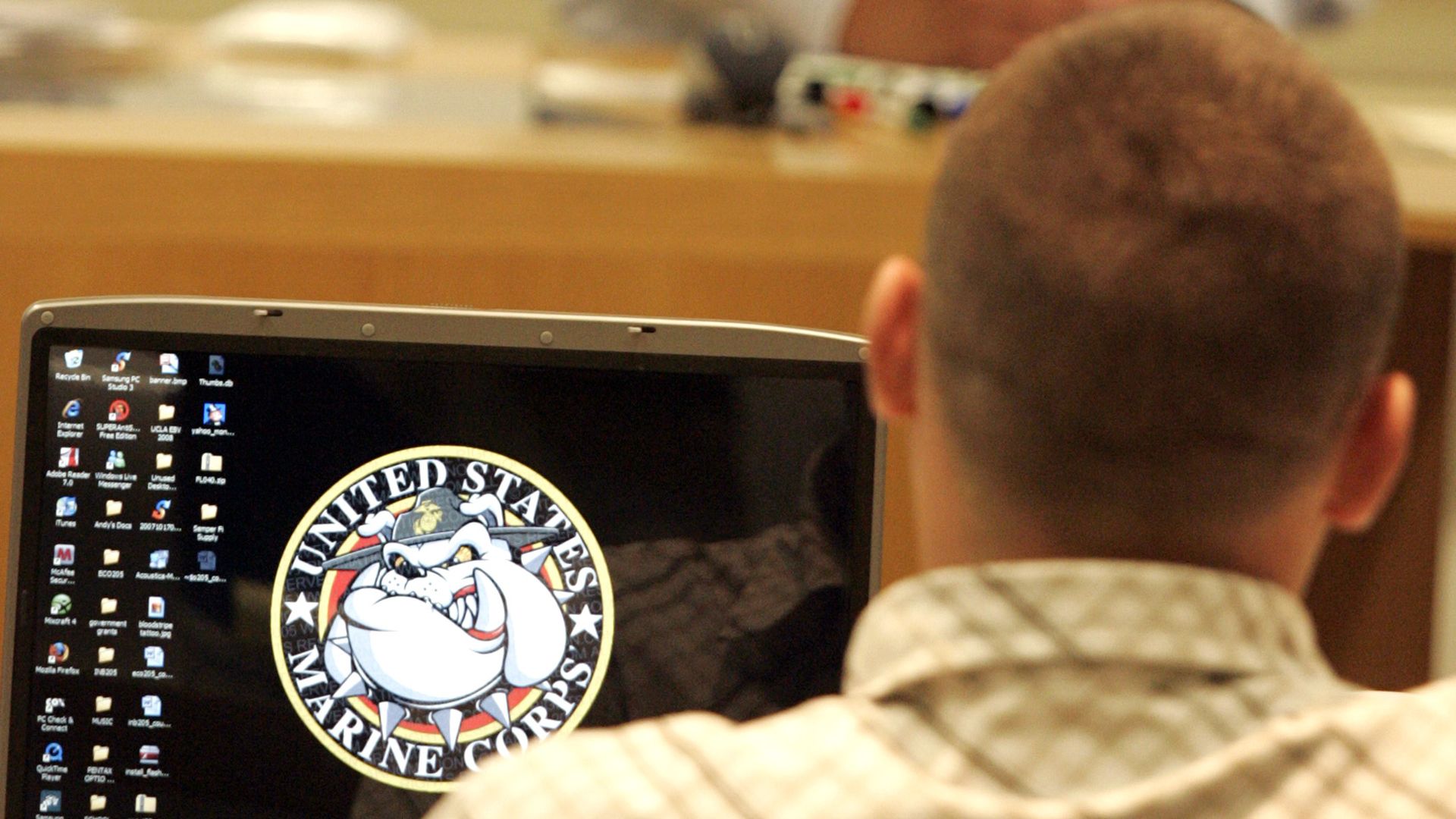 A man sits at a laptop displaying the US Marine Corps logo.