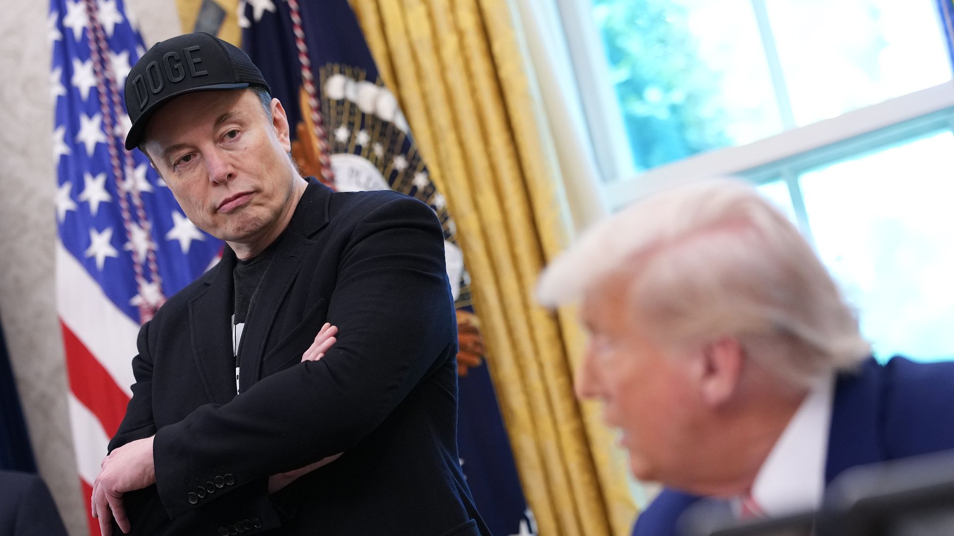 Elon Musk, in black jacket and black cap with "DOGE" stands with arms crossed in front of U.S. flag and presidential seal, looking at President Trump in blue suit partially visible.