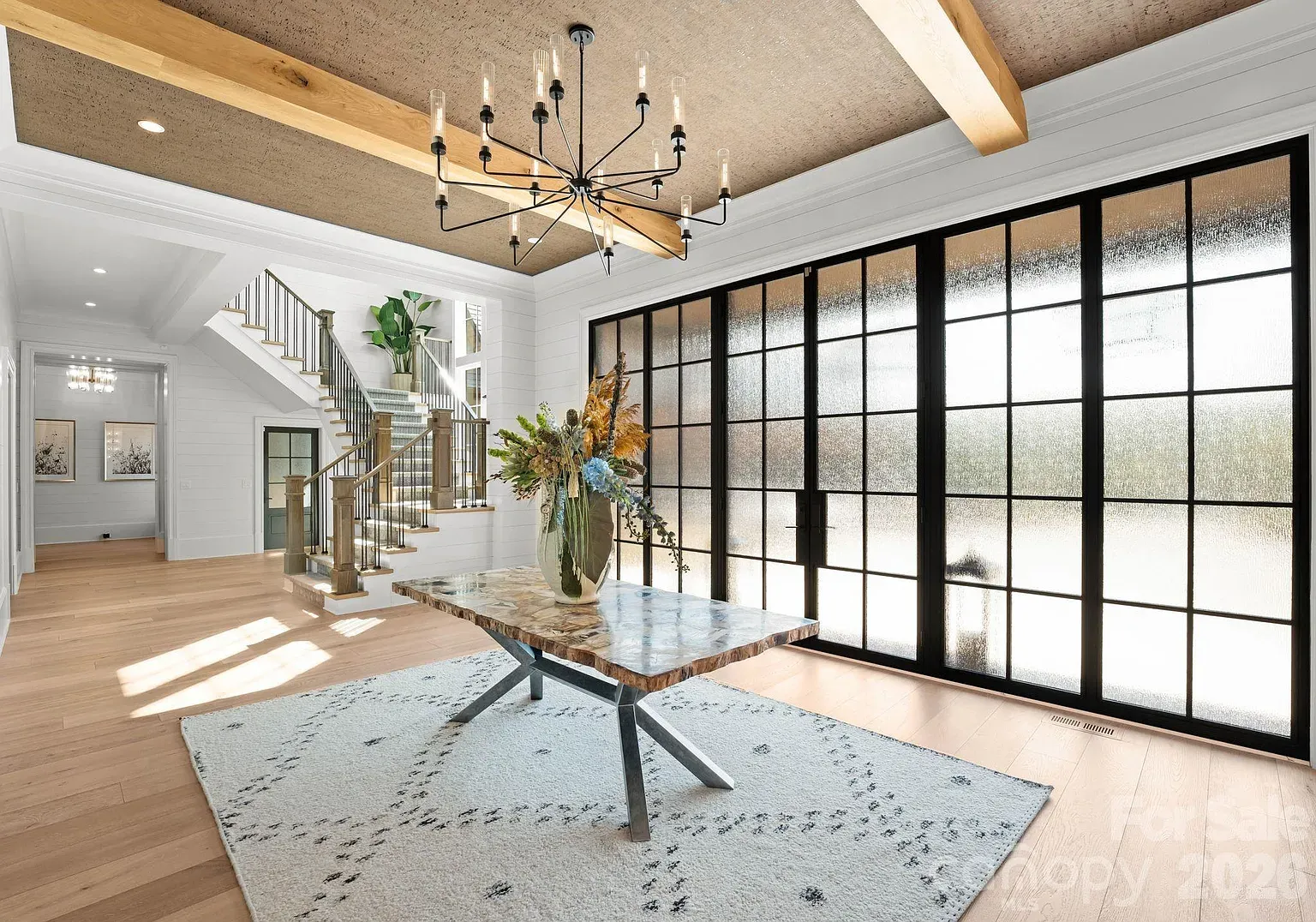 Bright modern foyer with a marble-topped table on a light rug, a black metal chandelier, exposed wooden beams, and frosted glass grid doors along the wall; an open staircase leads upstairs.