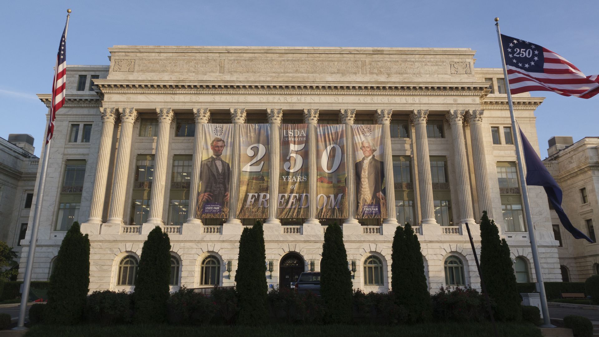 Grand neoclassical government building with tall columns. Banners advertising "250 Years of Freedom" hang across the front; US flags on both sides under a blue sky.
