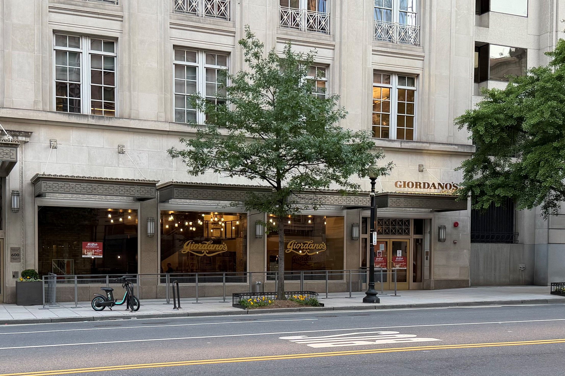 Urban street scene featuring a beige stone building with large windows and decorative balconies. A green tree frames Giordano's storefront; a teal electric scooter sits on the sidewalk.
