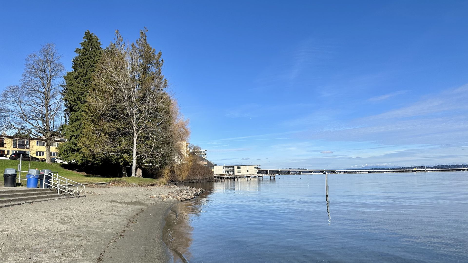 A grayish sandy beach with rocks and trees behind it and bridge over water in the background.