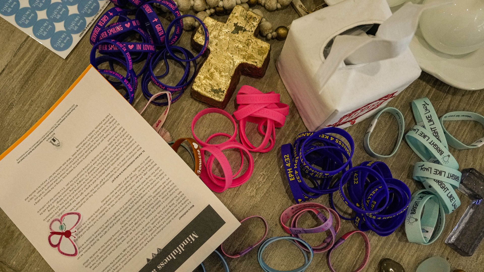 Colorful pink, blue and purple bracelets honoring campers who died in the Camp Mystic floods are seen sitting on a wooden table alongside a box of tissues and a golden cross.