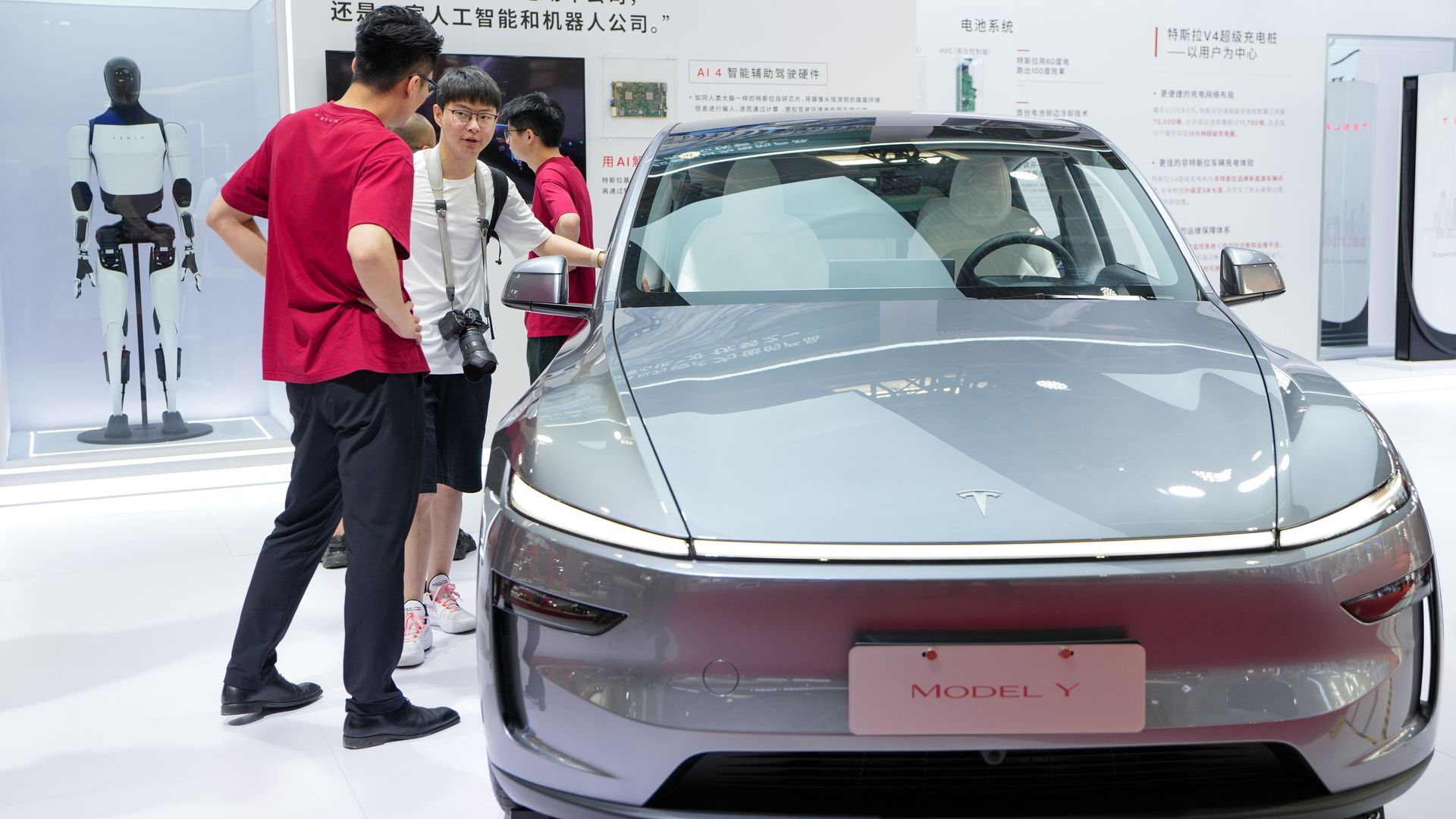 Three people talking beside a silver Tesla Model Y car inside a brightly lit exhibition space with Chinese text and a robot display labeled Tesla Bot in the background.