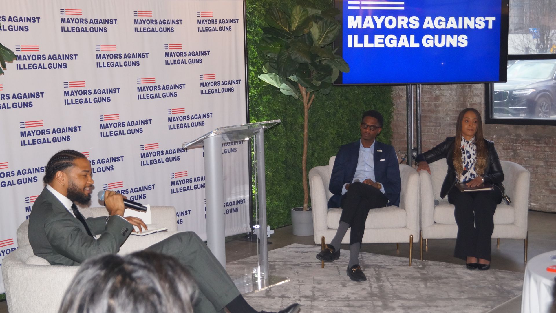 Man in chair with mic talks to two other people in white chairs under blue screen that says Mayors Against Illegal Guns