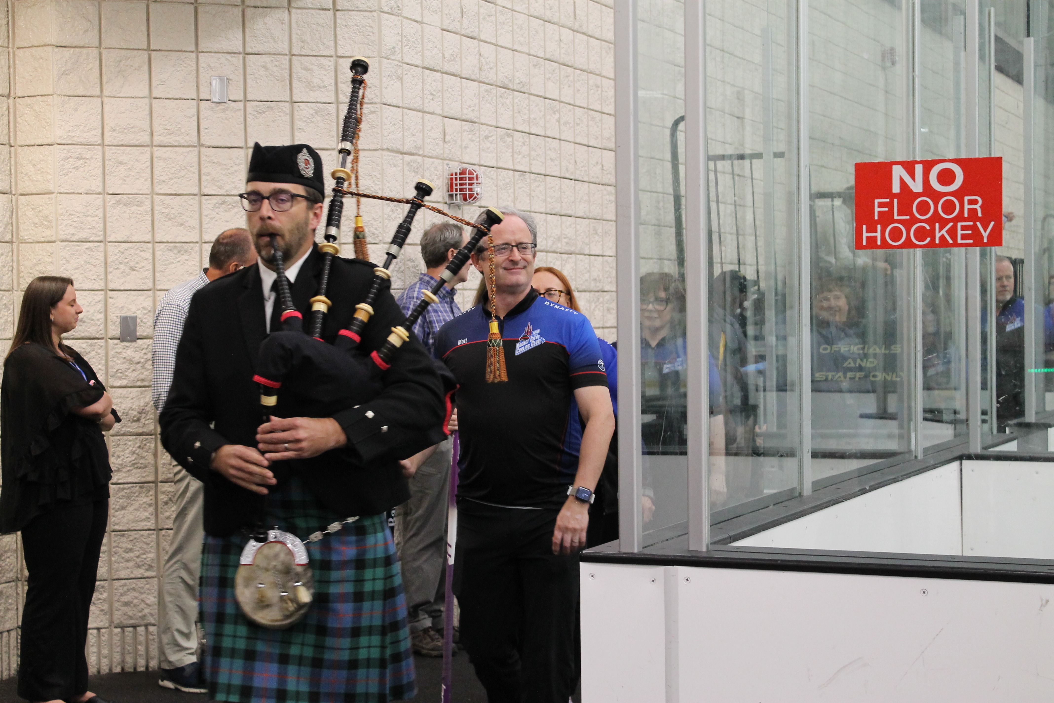Man in traditional Scottish attire playing bagpipes indoors near a wall, with several people standing behind him and a red sign reading "NO FLOOR HOCKEY" on glass panels.