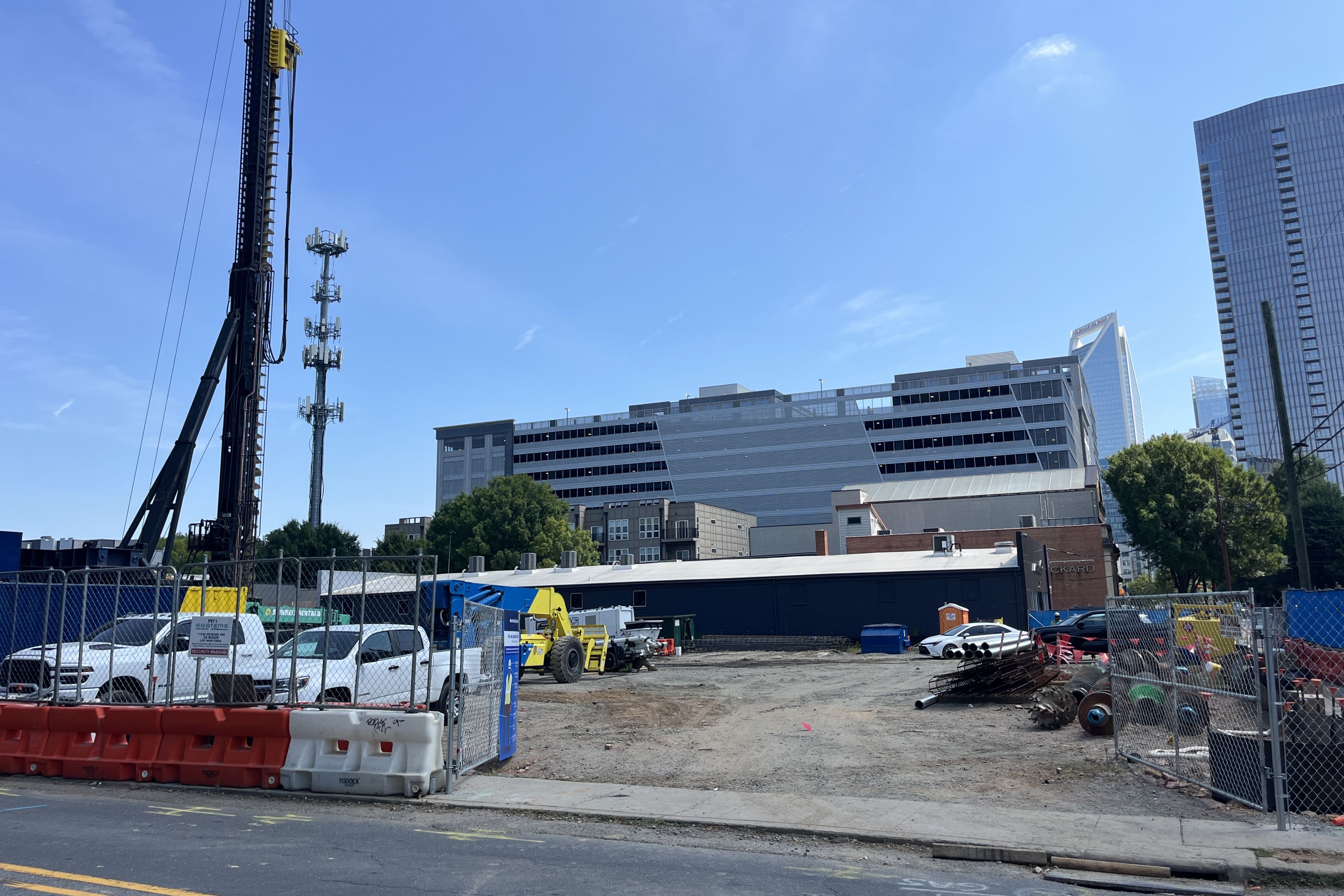 Construction site under clear blue sky. A tall crane/rig, fencing, yellow equipment, portable barriers in red and white at foreground. Modern buildings and a parking structure in background.