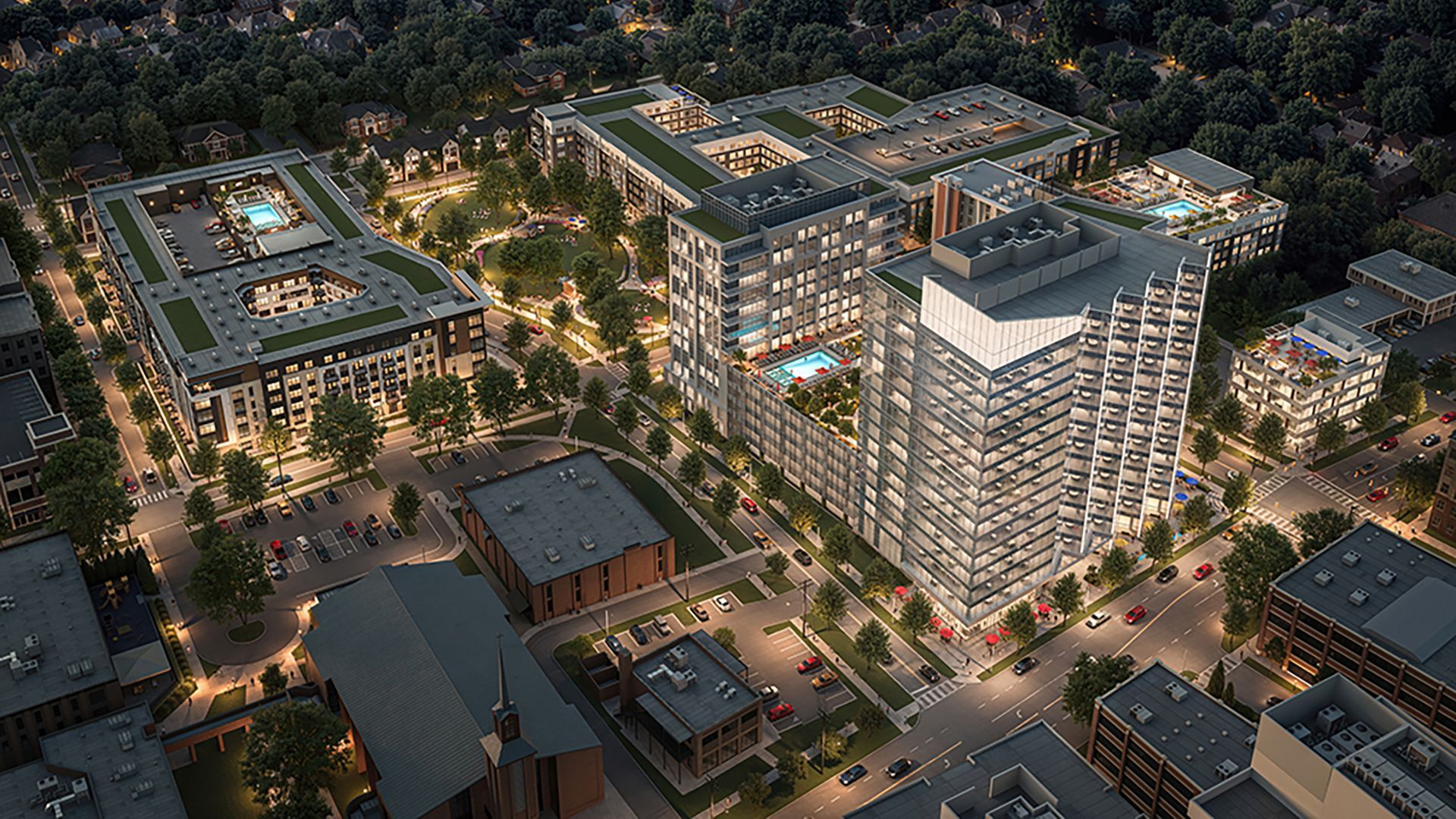 Night aerial view of a modern urban complex with lit multi-story buildings, rooftop pools, tree-lined streets, parking lots, and a central green space with pathways and trees.