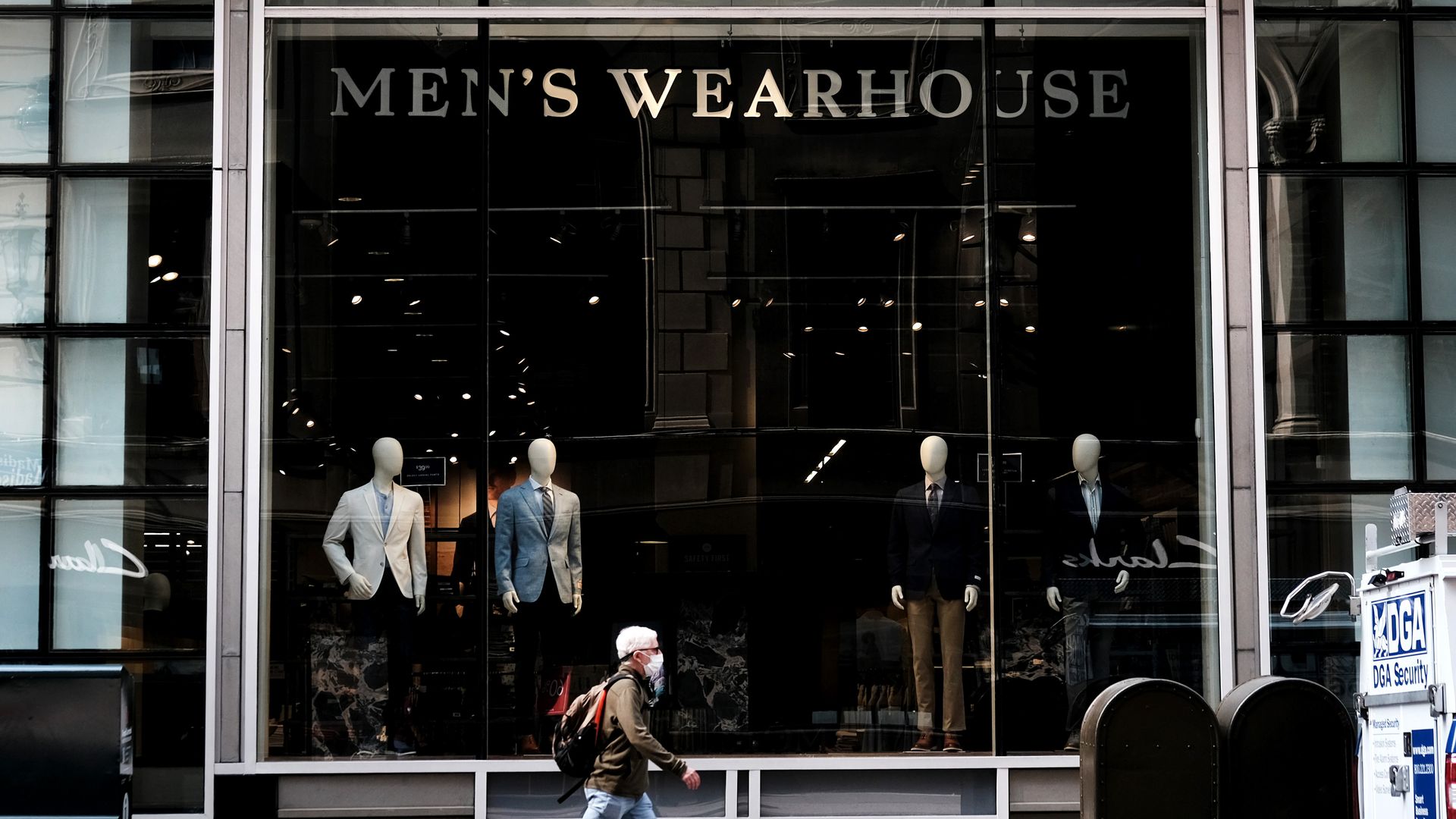 A pedestrian walks in front of a Men's Wearhouse store facade, with the signage spelled out in gold lettering, located on an urban city street.