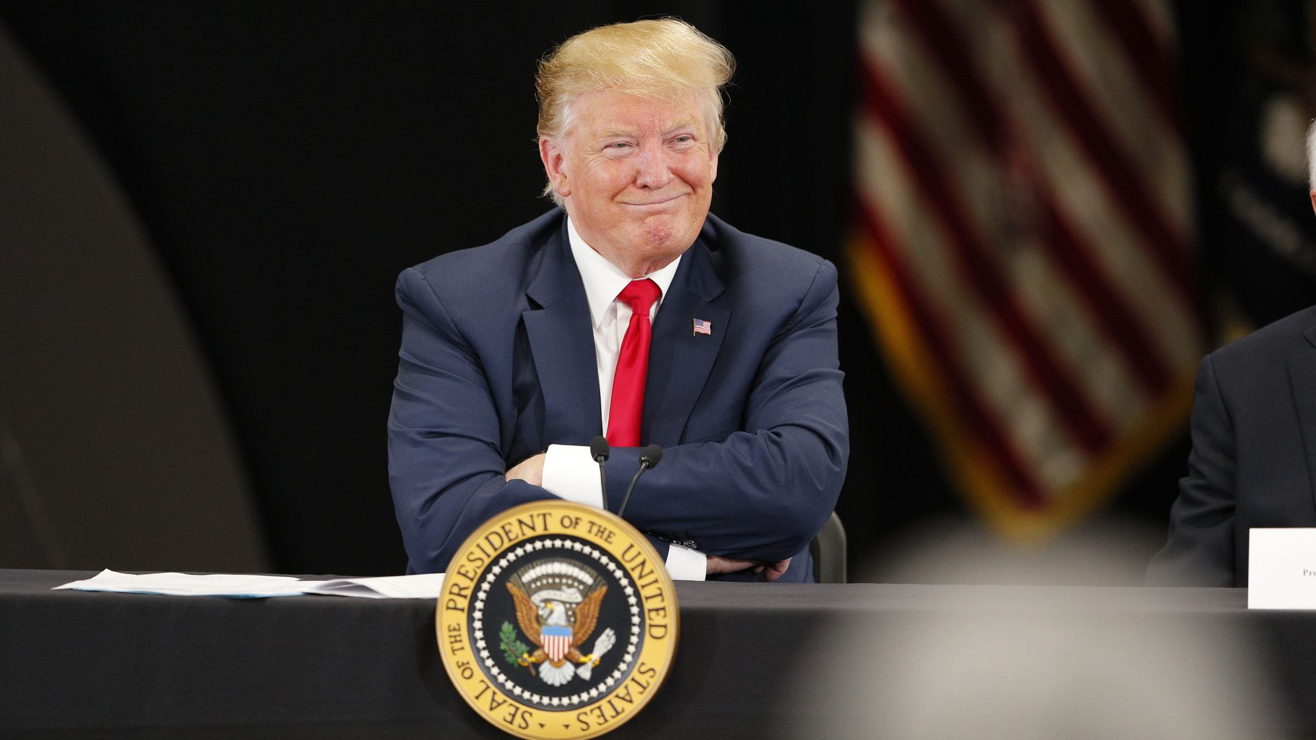 In this image, Trump sits and smiles with his arms folded with the American flag next to him.