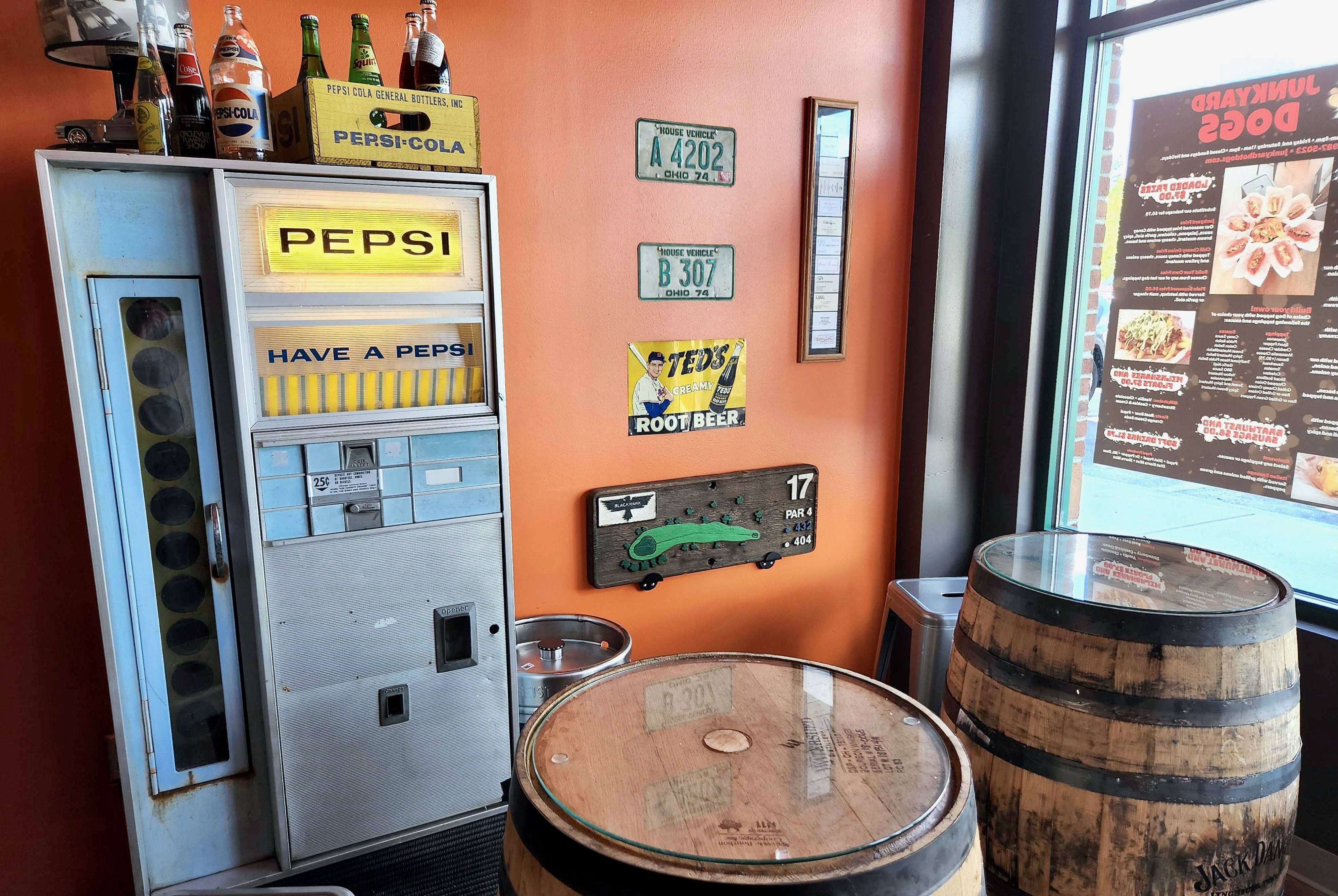 A vintage Pepsi vending machine amid two large barrels and old license plates lining an orange wall