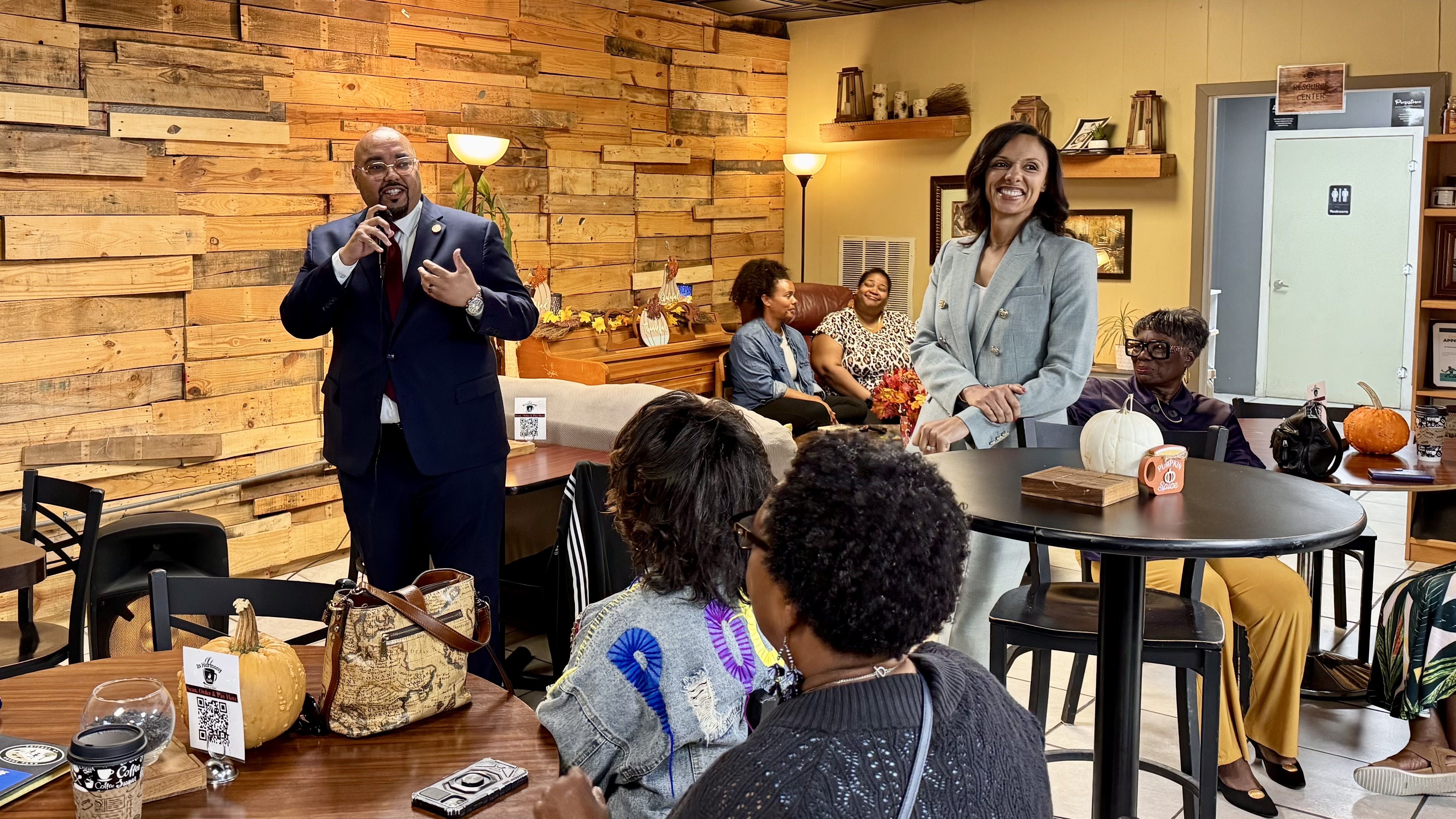 Durhal speaks into a microphone on the left. Sheffield is nearby, smiling, as Durhal speaks. They are in front of a small audience
