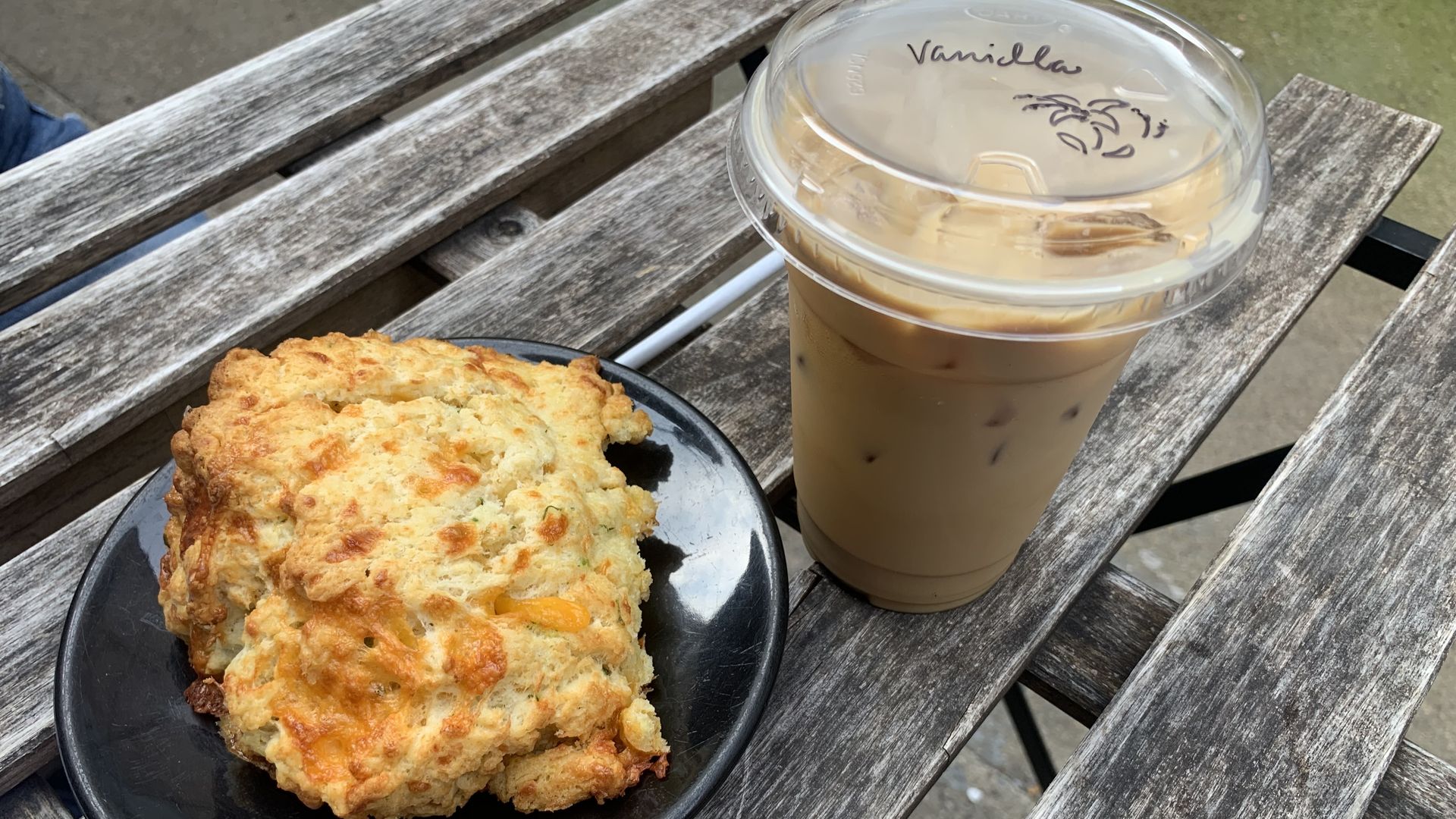 A golden-brown cheese biscuit on a black plate next to a plastic cup of iced vanilla coffee with a clear lid, both placed on a weathered wooden outdoor table.