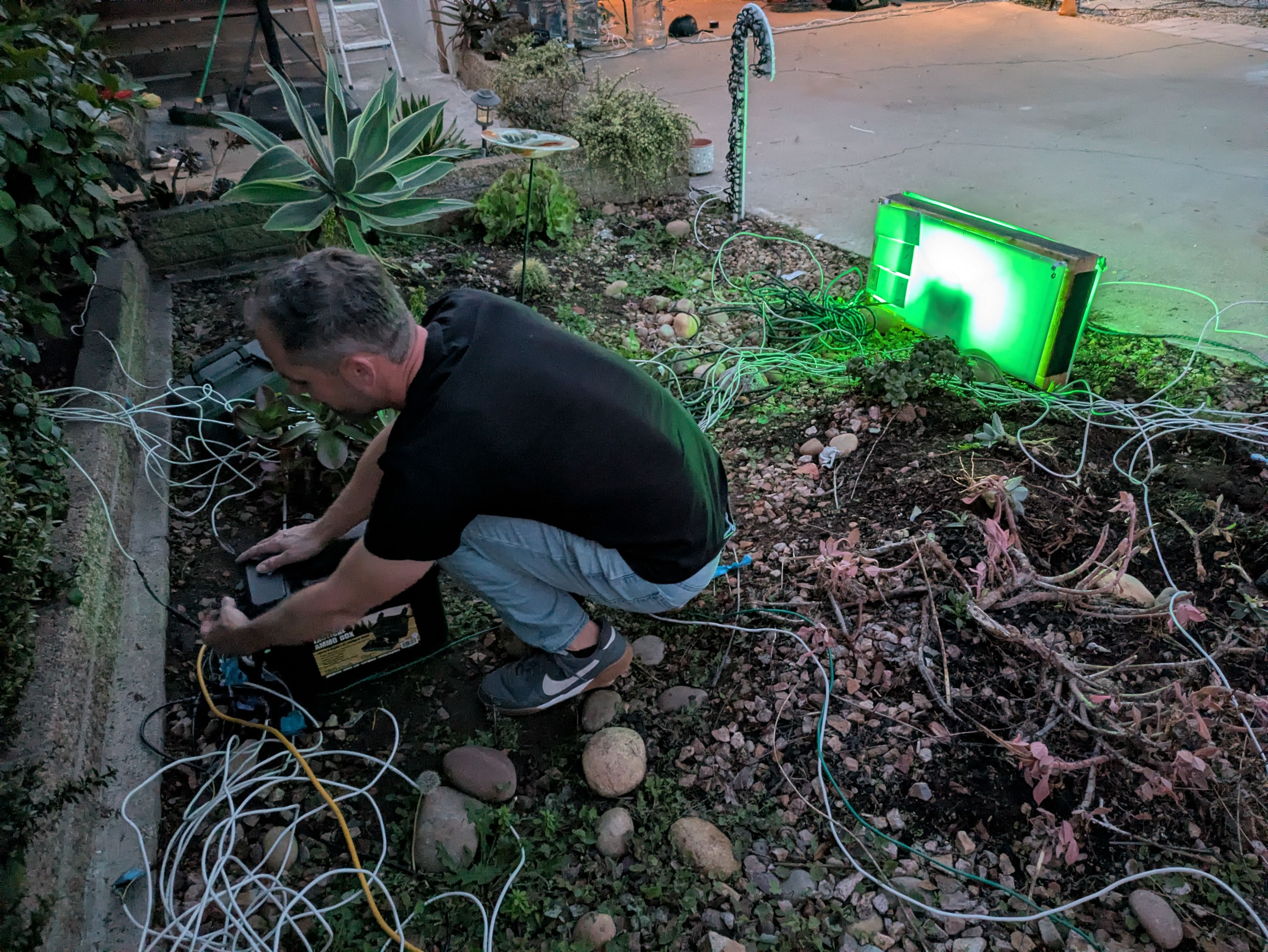 Man in black shirt and jeans kneeling in garden at dusk, handling a black box with many tangled wires around. Green light glows from rectangular device nearby.