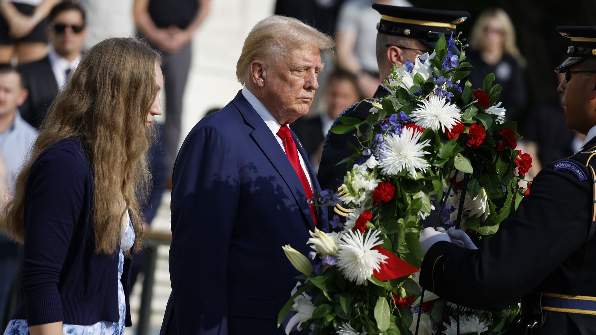 Donald Trump, wearing a blue suit, a red tie and a white shirt, stands in front of a wreath held by a soldier.